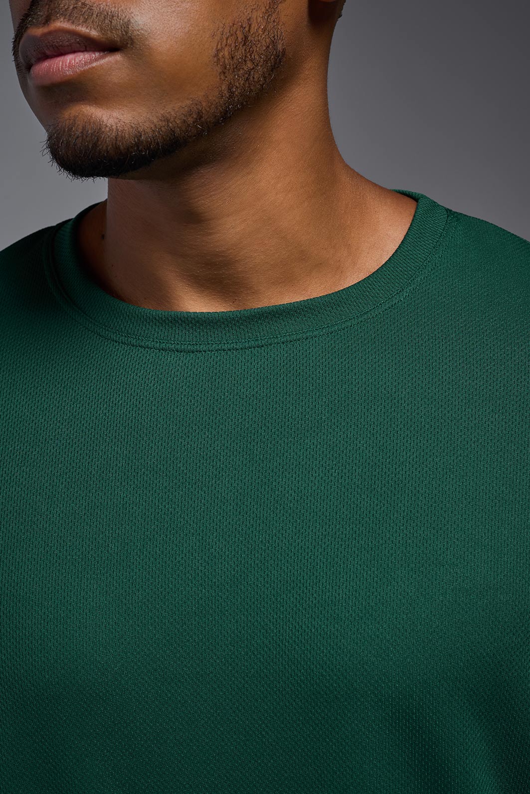 A male model wearing the men's Oxymesh Long Sleeve Tech Tee in the color forest green, made out of 100% polyester, showing the relaxed fit from a close-up collar view. This shirt is breathable, quick-drying, moisture-wicking, and made in the USA. #forest-green