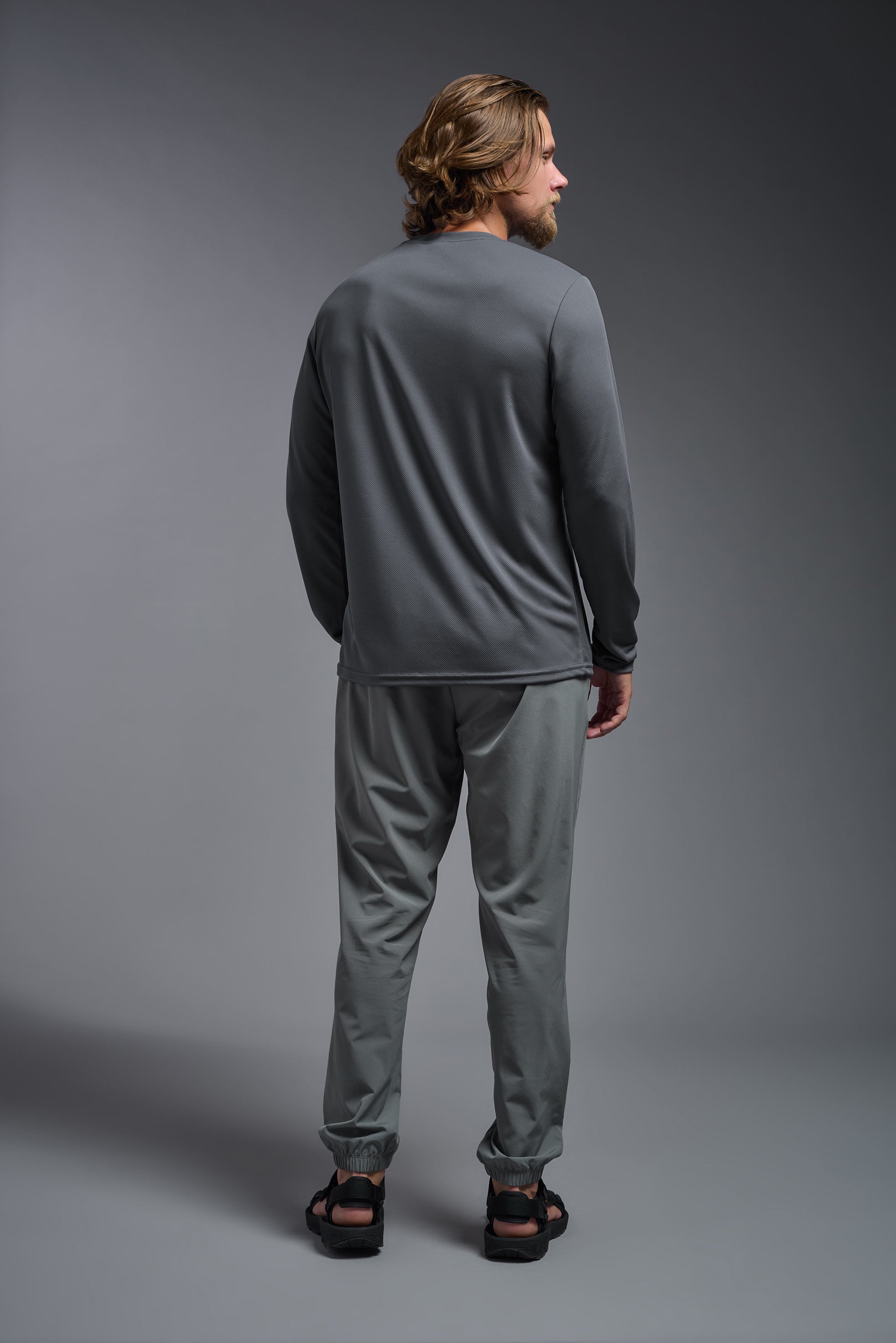 A male model wearing the men's Oxymesh Long Sleeve Tech Tee in the color graphite gray made out of 100% polyester, showing the relaxed fit from a back view. This shirt is breathable, quick-drying, moisture-wicking, and made in the USA. #graphite-gray