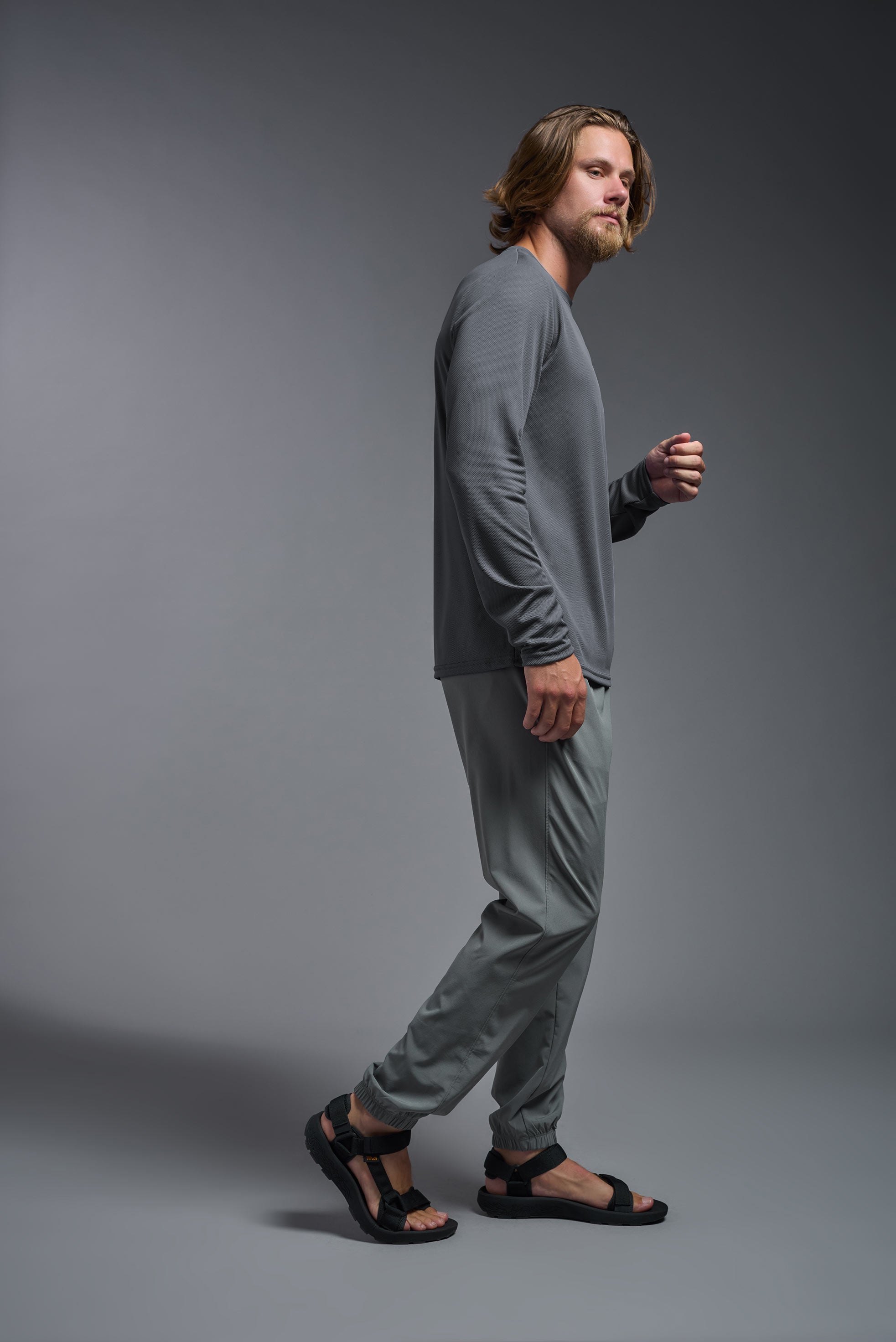 A male model wearing the men's Oxymesh Long Sleeve Tech Tee in the color graphite gray made out of 100% polyester, showing the relaxed fit from a front side view. This shirt is breathable, quick-drying, moisture-wicking, and made in the USA. #graphite-gray