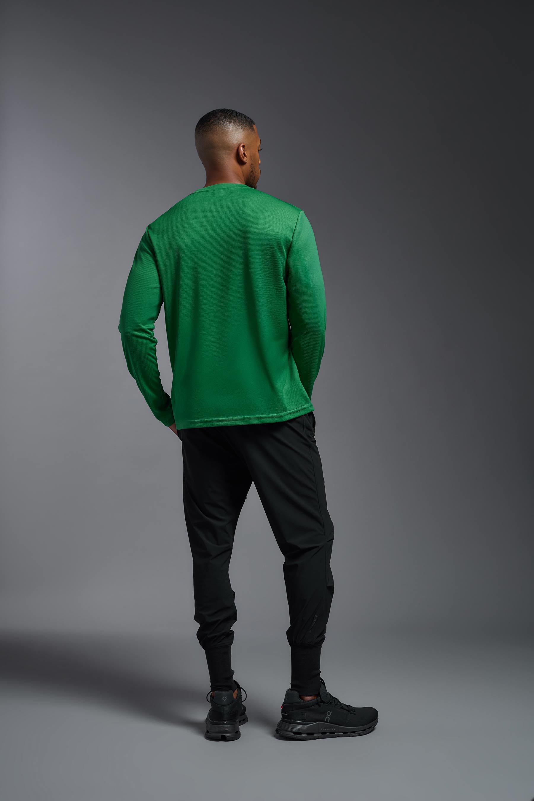 A male model wearing the men's Oxymesh Long Sleeve Tech Tee in the color kelly green, made out of 100% polyester, showing the relaxed fit from a back view. This shirt is breathable, quick-drying, moisture-wicking, and made in the USA. #kelly-green