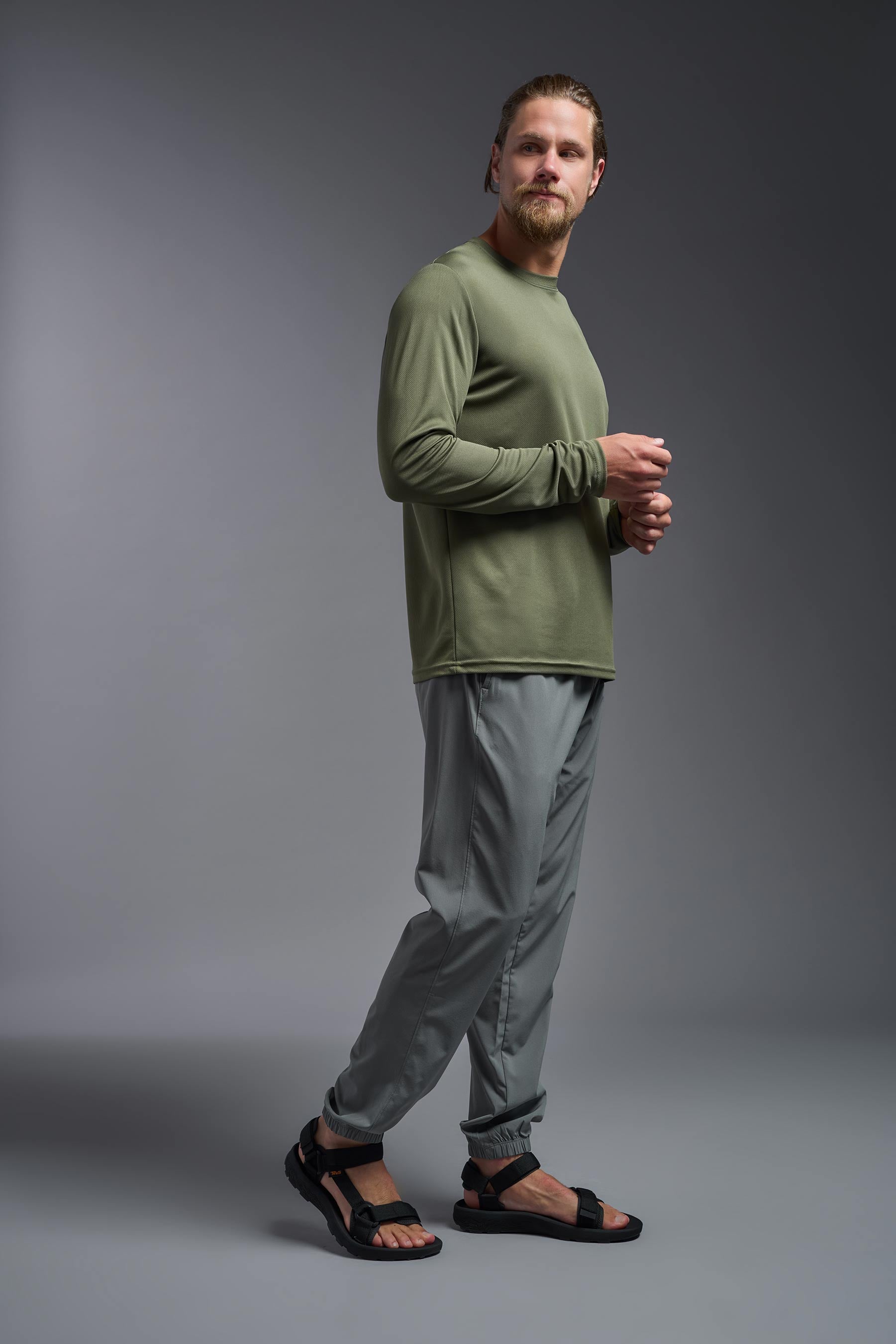 A male model wearing the men's Oxymesh Long Sleeve Tech Tee in the color military green, made out of 100% polyester, showing the relaxed fit from a side view. This shirt is breathable, quick-drying, moisture-wicking, and made in the USA. #military-green
