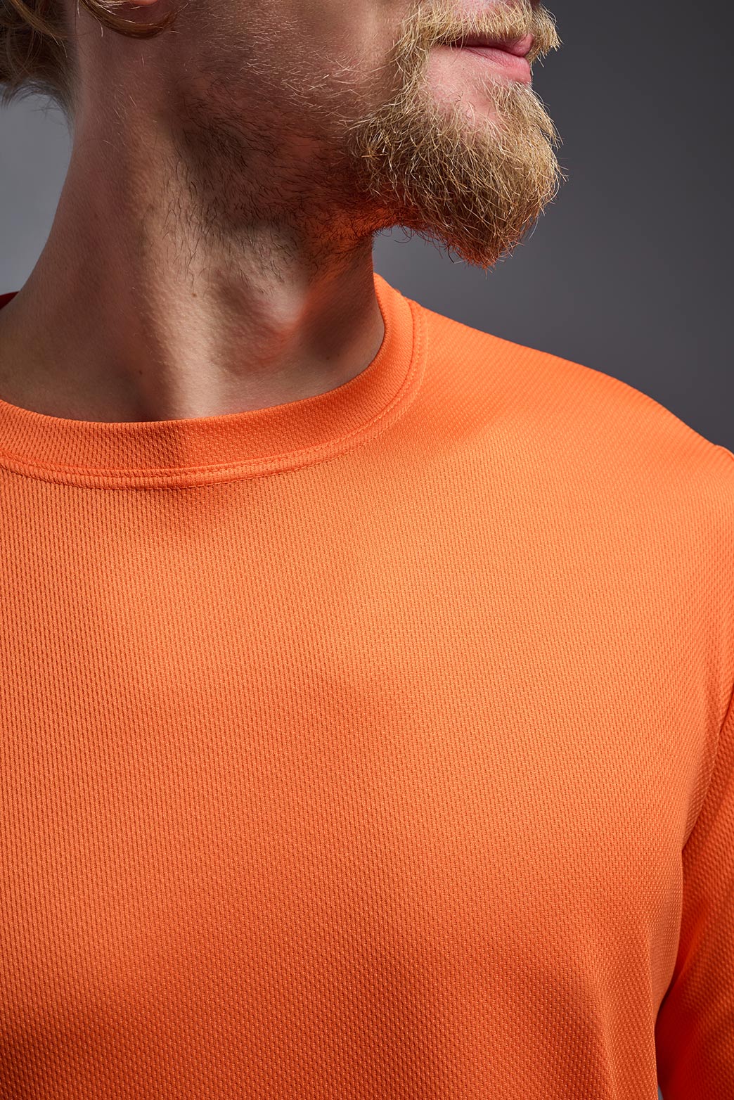 A male model wearing the men's Oxymesh Long Sleeve Tech Tee in the color orange made out of 100% polyester, showing the relaxed fit from a close-up collar view. This shirt is breathable, quick-drying, moisture-wicking, and made in the USA. #orange
