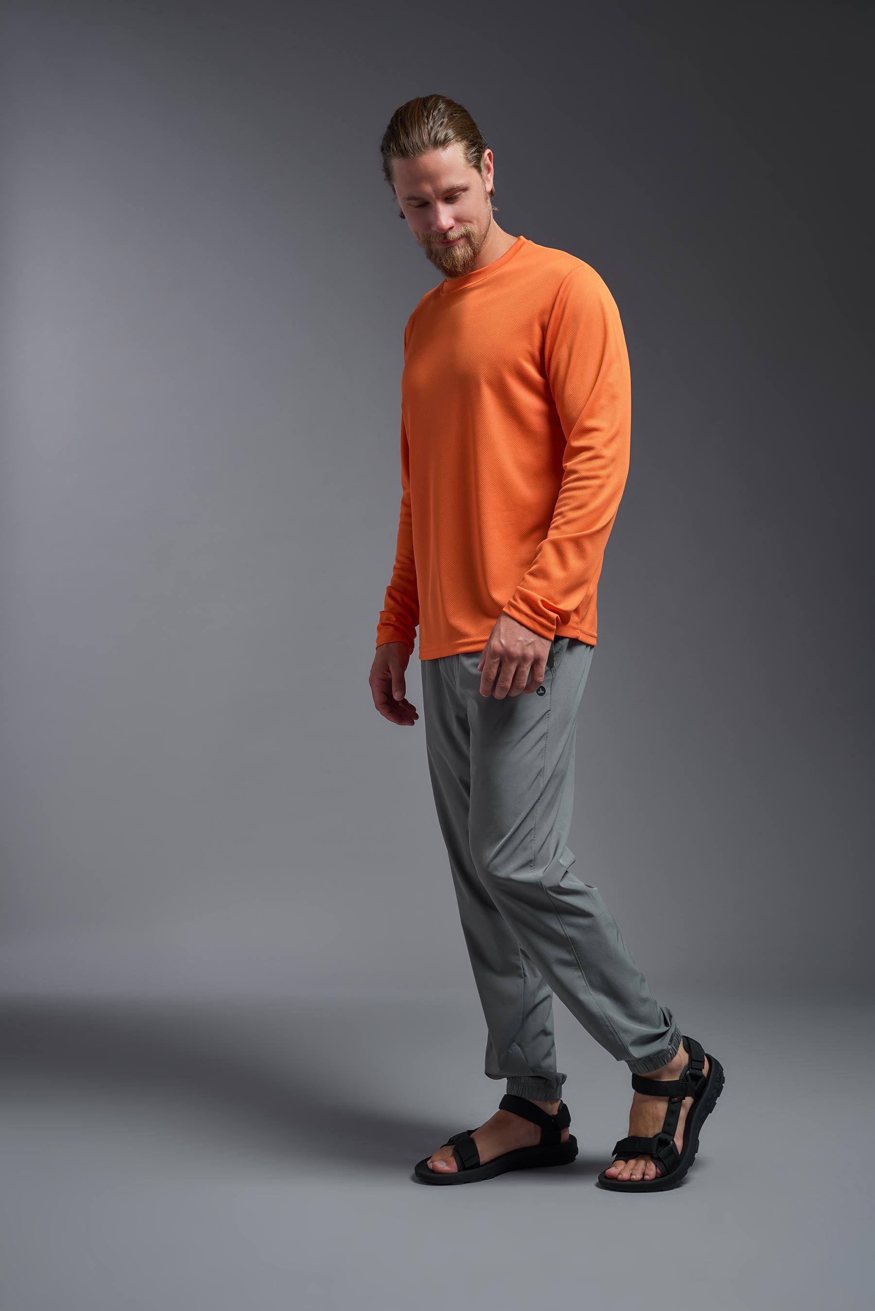 A male model wearing the men's Oxymesh Long Sleeve Tech Tee in the color orange made out of 100% polyester, showing the relaxed fit from a side view. This shirt is breathable, quick-drying, moisture-wicking, and made in the USA. #orange