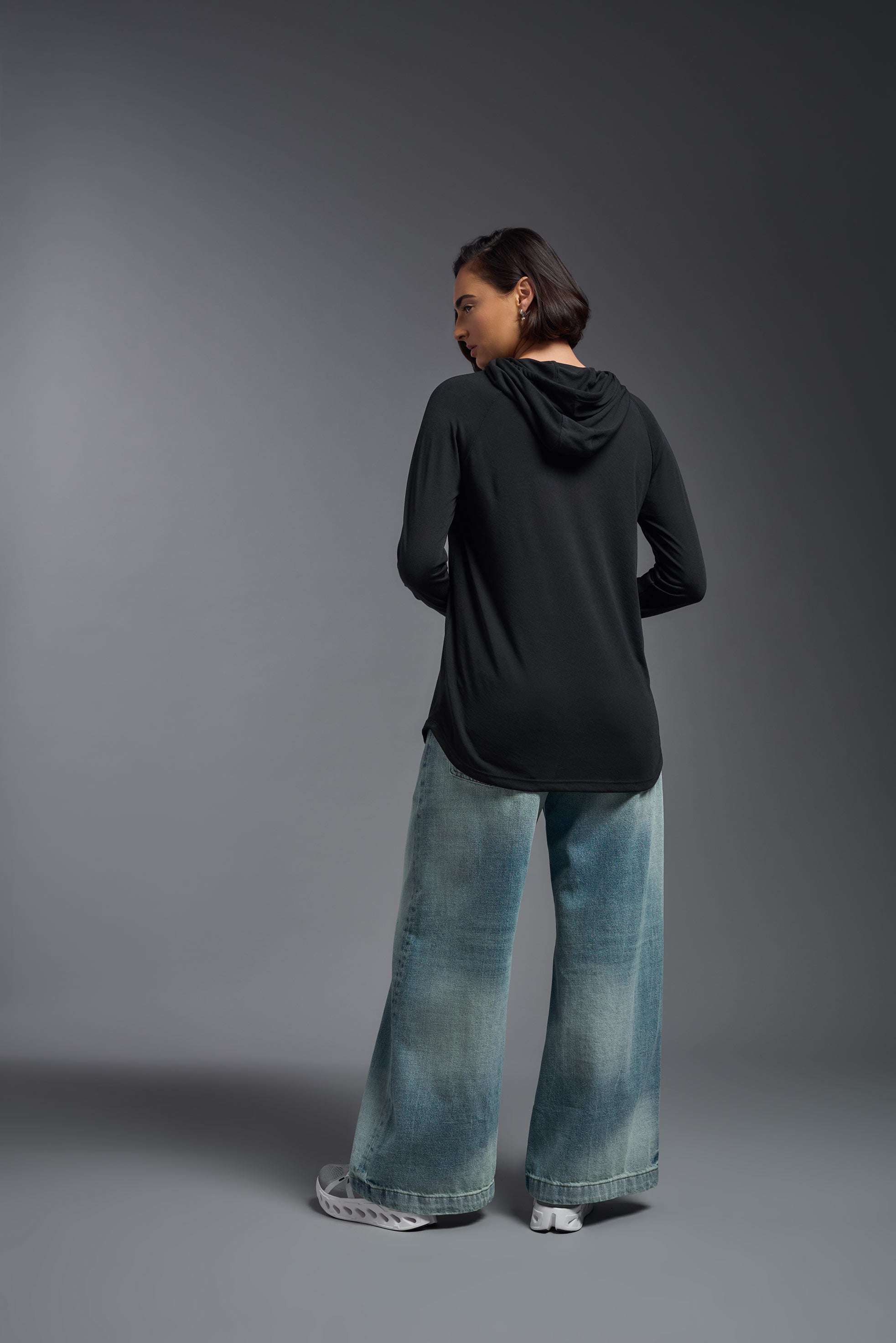 A female model wearing the women's Siro™ Hoodie Shirt in the color black, made out of 100% filament polyester, showing the relaxed fit from a back view. This long sleeve hoodie shirt is breathable, quick-drying, moisture-wicking, and made in the USA. #black