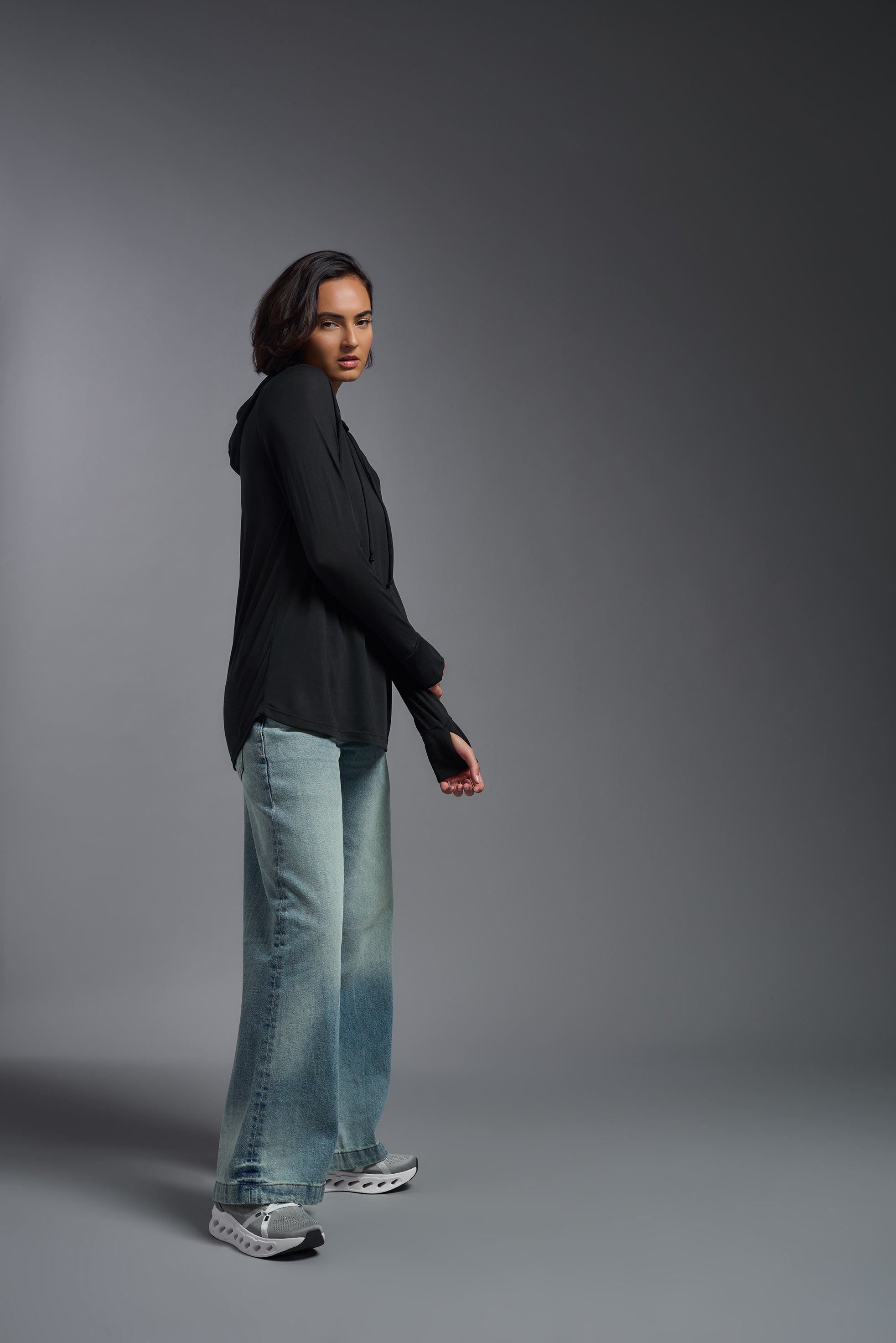 A female model wearing the women's Siro™ Hoodie Shirt in the color black, made out of 100% filament polyester, showing the relaxed fit from a side view. This long sleeve hoodie shirt is breathable, quick-drying, moisture-wicking, and made in the USA. #black