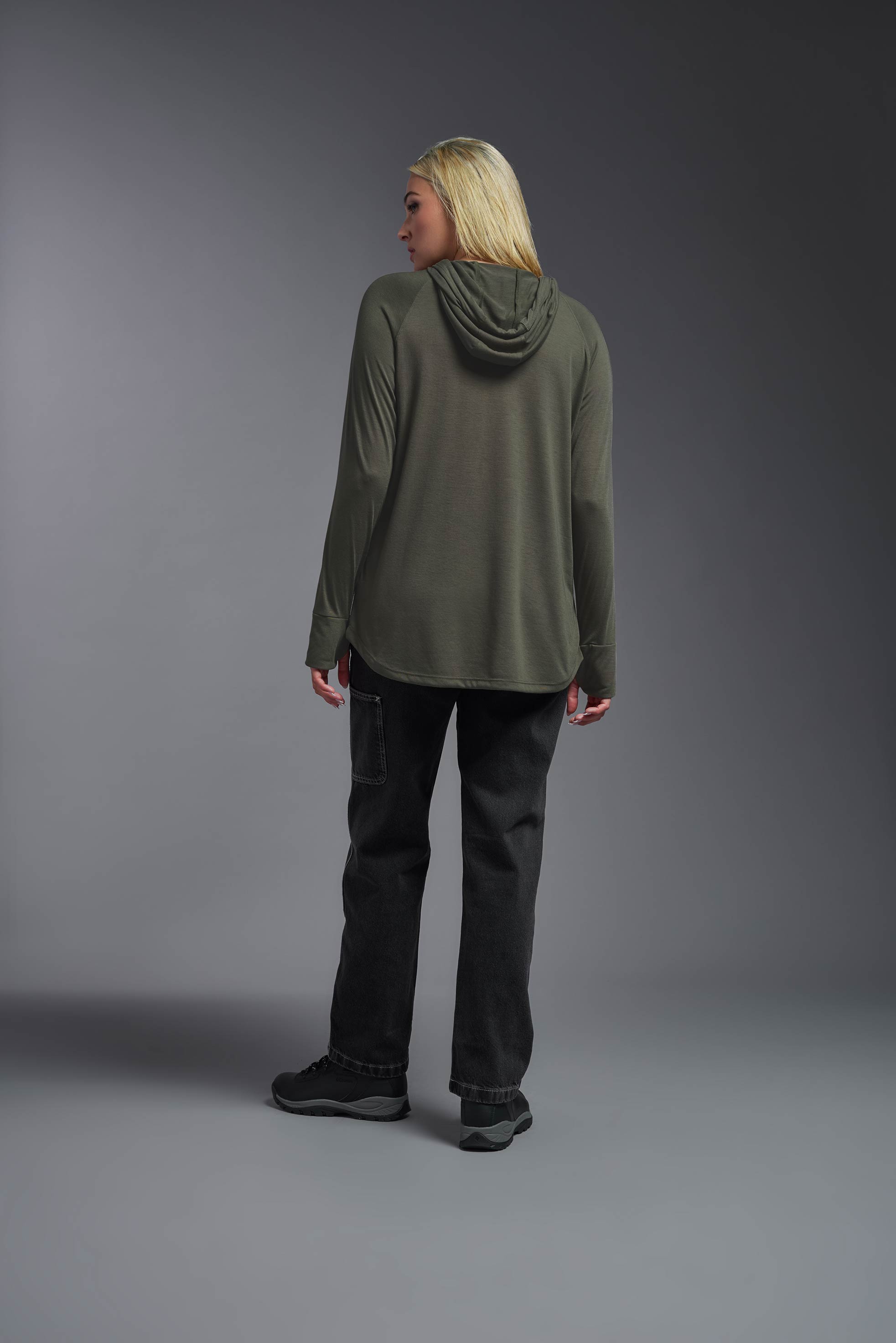 A female model wearing the women's Siro™ Hoodie Shirt in the color olive, made out of 100% filament polyester, showing the relaxed fit from a full body back view. This long sleeve hoodie shirt is breathable, quick-drying, moisture-wicking, and made in the USA. #olive