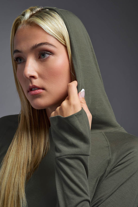 A female model wearing the women's Siro™ Hoodie Shirt in the color olive, made out of 100% filament polyester, showing the relaxed fit from a front closeup hood detail view. This long sleeve hoodie shirt is breathable, quick-drying, moisture-wicking, and made in the USA. #olive