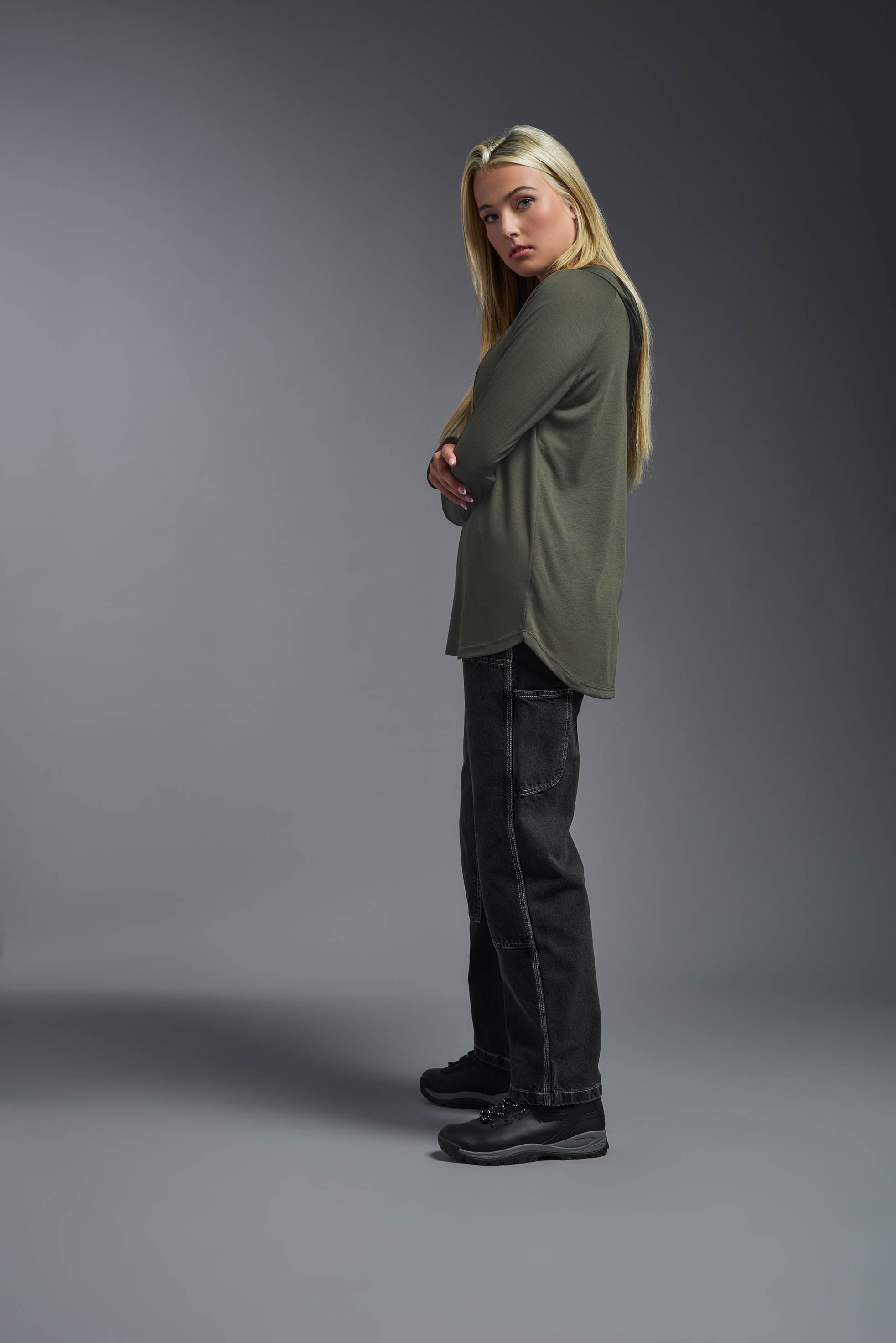 A female model wearing the women's Siro™ Hoodie Shirt in the color olive, made out of 100% filament polyester, showing the relaxed fit from a side view. This long sleeve hoodie shirt is breathable, quick-drying, moisture-wicking, and made in the USA. #olive