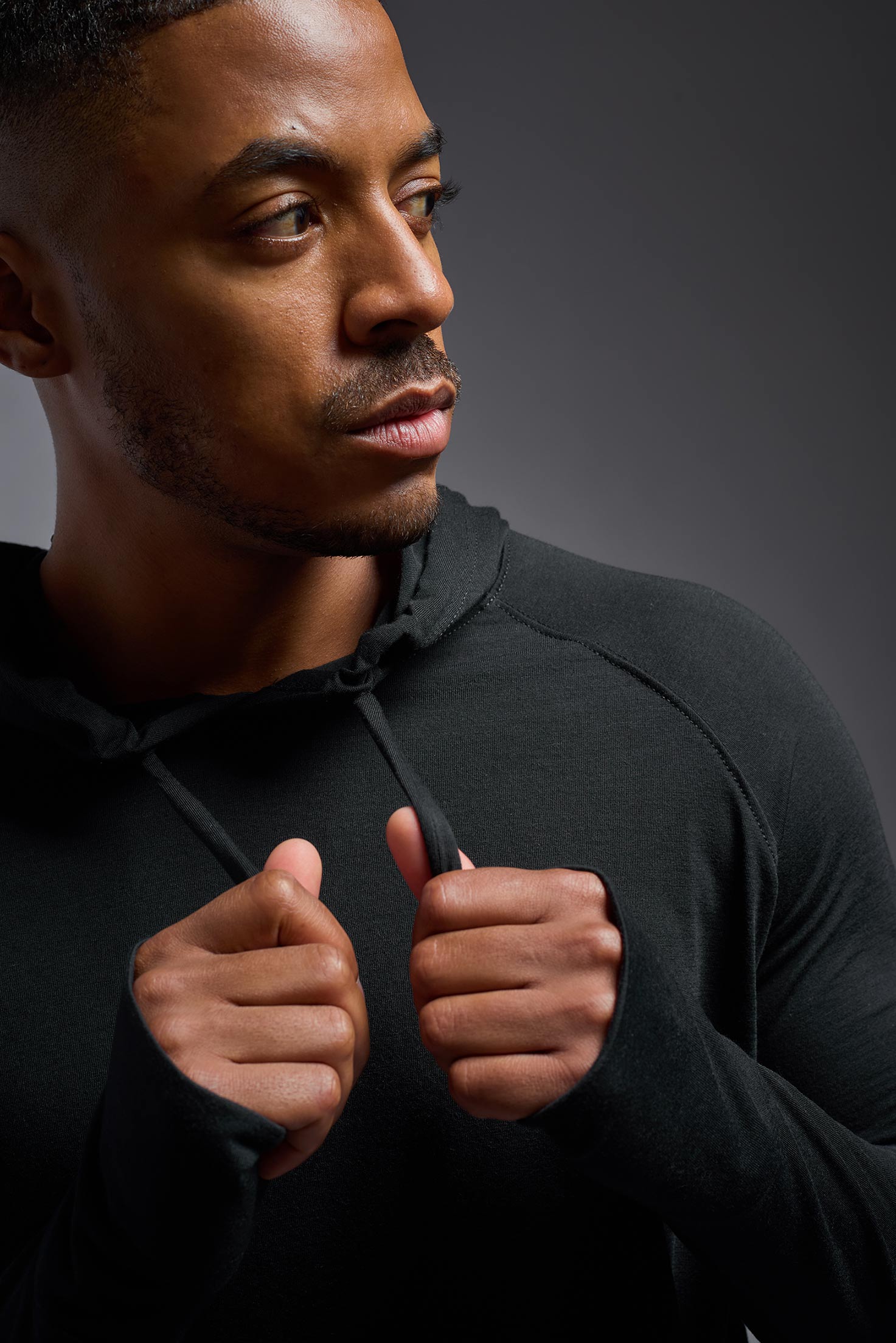 A male model wearing the men's Siro™ Hoodie Shirt in the color black, made out of 100% polyester, showing the relaxed fit from a detail view. This long sleeve hoodie shirt is breathable, quick-drying, moisture-wicking, and made in the USA. #black