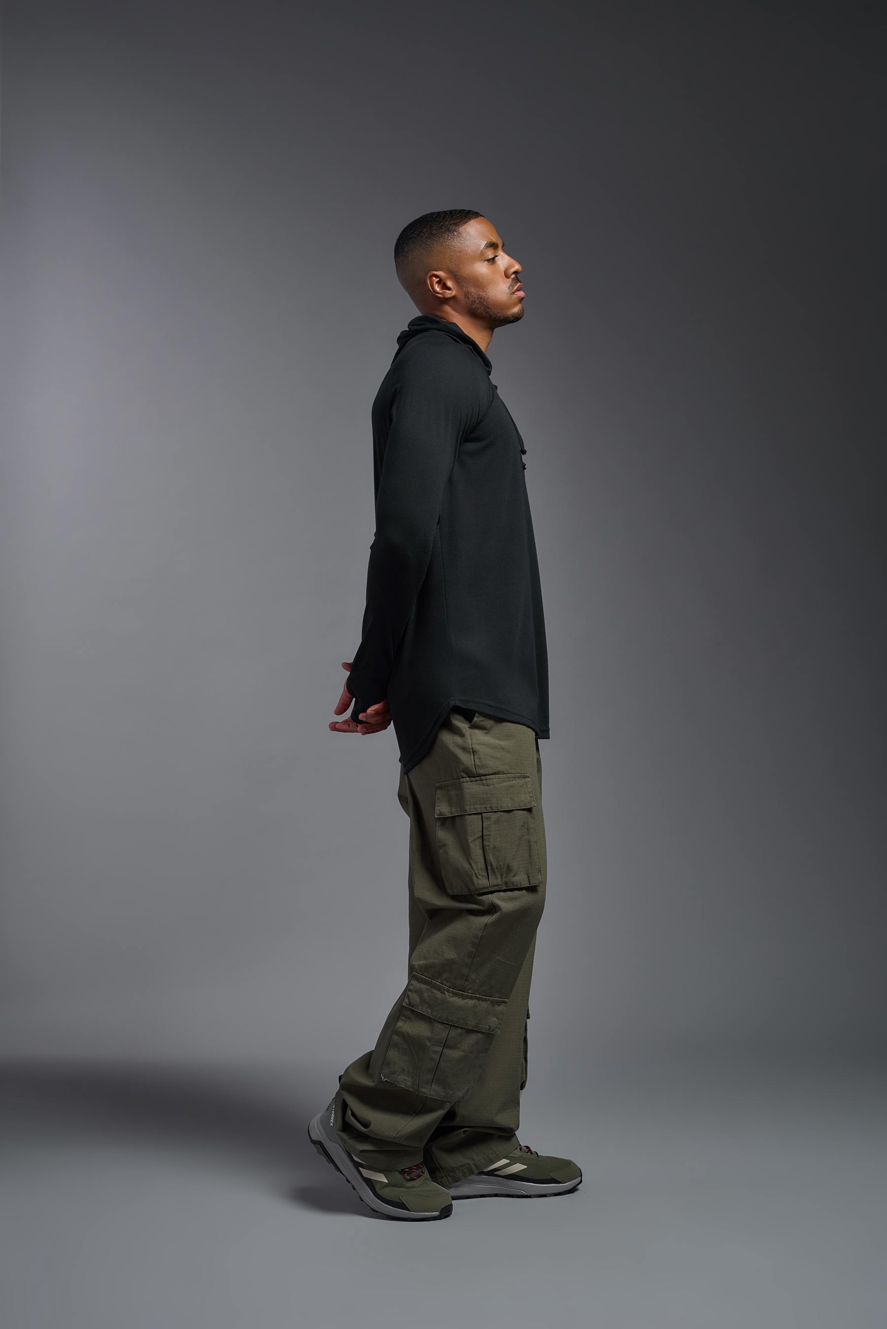 A male model wearing the men's Siro™ Hoodie Shirt in the color black, made out of 100% polyester, showing the relaxed fit from a side view. This long sleeve hoodie shirt is breathable, quick-drying, moisture-wicking, and made in the USA. #black