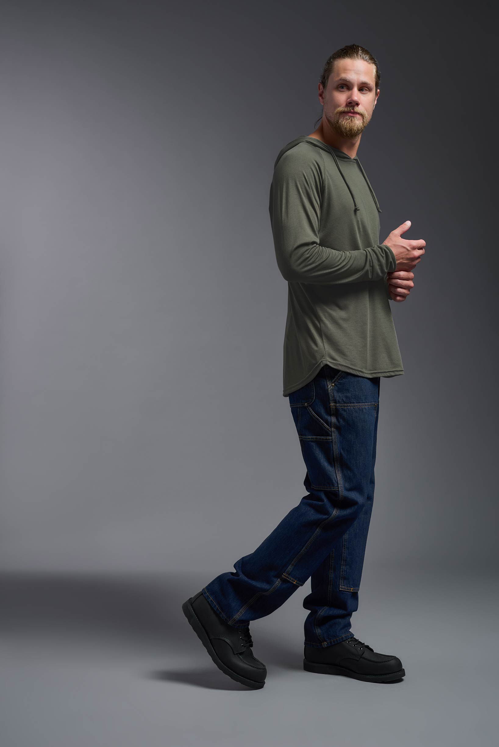 A male model wearing the men's Siro™ Hoodie Shirt in the color olive, made out of 100% polyester, showing the relaxed fit from a side view. This long sleeve hoodie shirt is breathable, quick-drying, moisture-wicking, and made in the USA. #olive