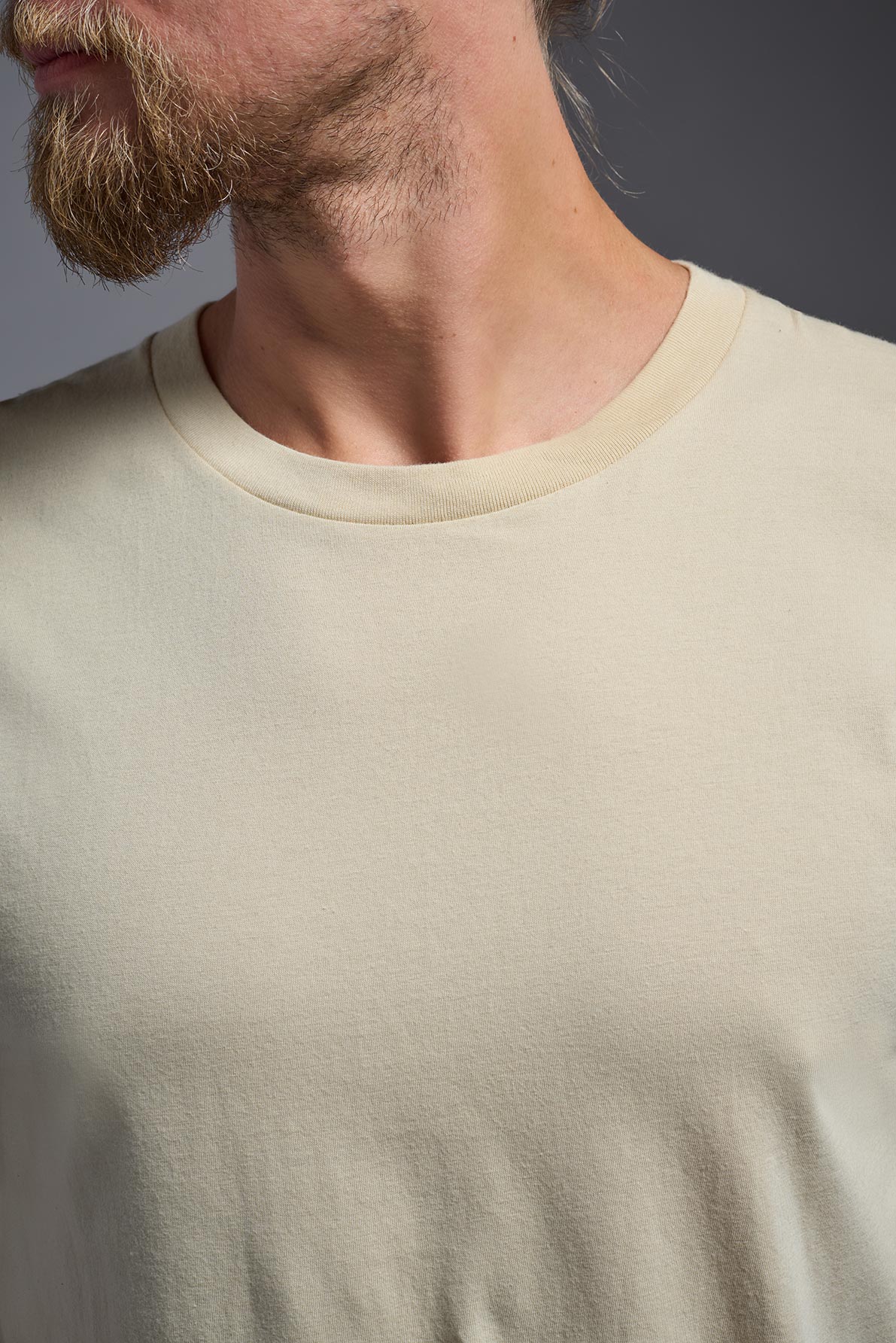 A male model wearing the unisex Americana Cotton Soft Tee in the color sand, made from 100% combed ring-spun USA-grown cotton, showing the classic fit from a close-up collar view. This premium shirt is Made in the USA. #sand