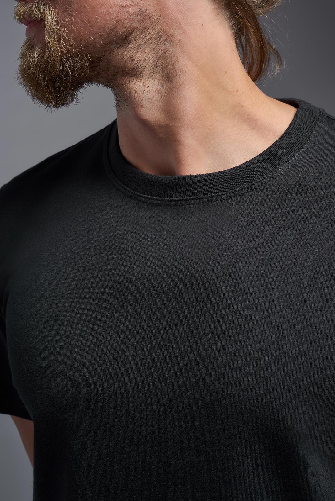 A male model wearing the unisex Imagen Short Sleeve Crewneck T-Shirt in the color black, made from 50% U.S. Grown Regenerative Cotton and 50% Recycled Polyester, showing the Relaxed Fit from a close-up collar view. This sustainable shirt is Made in the USA. #black