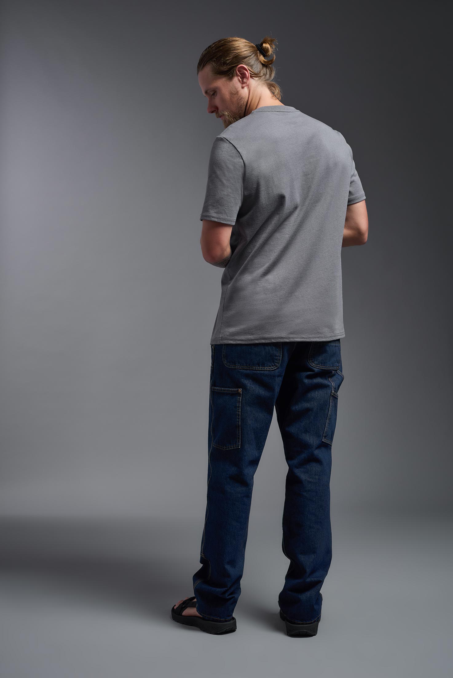 A male model wearing the unisex Imagen Short Sleeve Crewneck T-Shirt in the color carbon, made from 50% U.S. Grown Regenerative Cotton and 50% Recycled Polyester, showing the Relaxed Fit from a back view. This sustainable shirt is Made in the USA. #carbon