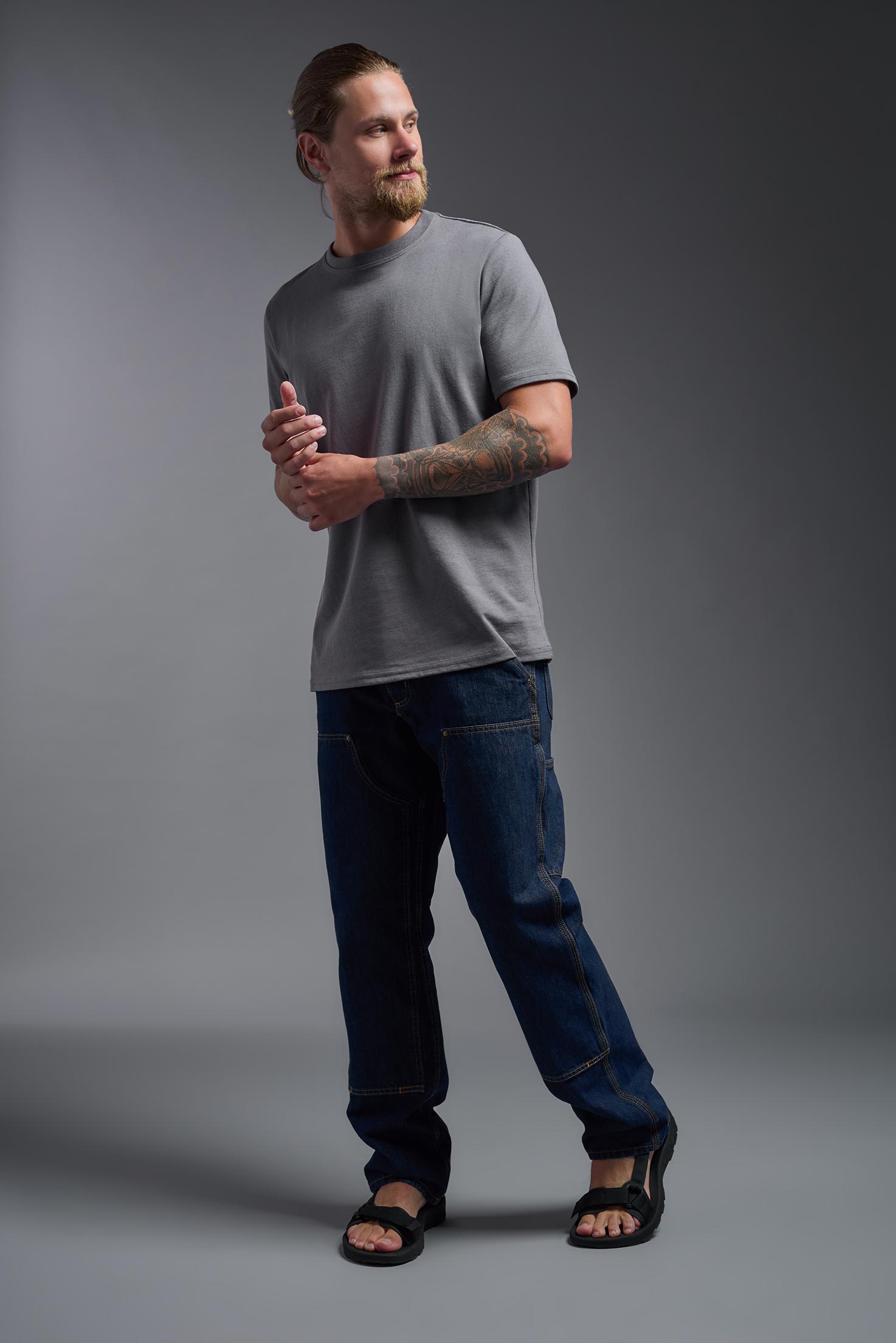 A male model wearing the unisex Imagen Short Sleeve Crewneck T-Shirt in the color carbon, made from 50% U.S. Grown Regenerative Cotton and 50% Recycled Polyester, showing the Relaxed Fit from a side view. This sustainable shirt is Made in the USA. #carbon