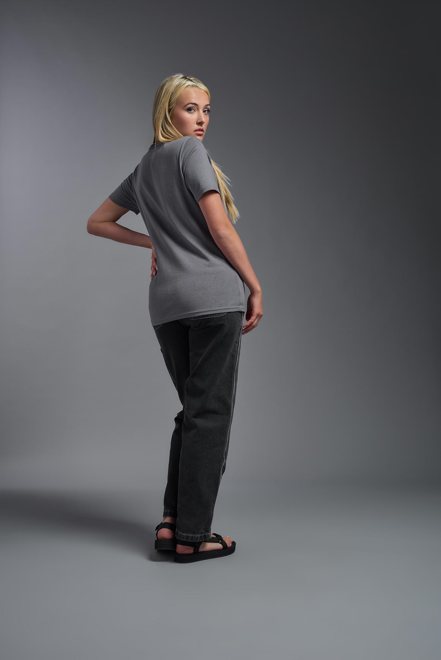 A female model wearing the unisex Imagen Short Sleeve Crewneck T-Shirt in the color carbon, made from 50% U.S. Grown Regenerative Cotton and 50% Recycled Polyester, showing the Relaxed Fit from a back view. This sustainable shirt is Made in the USA. #carbon
