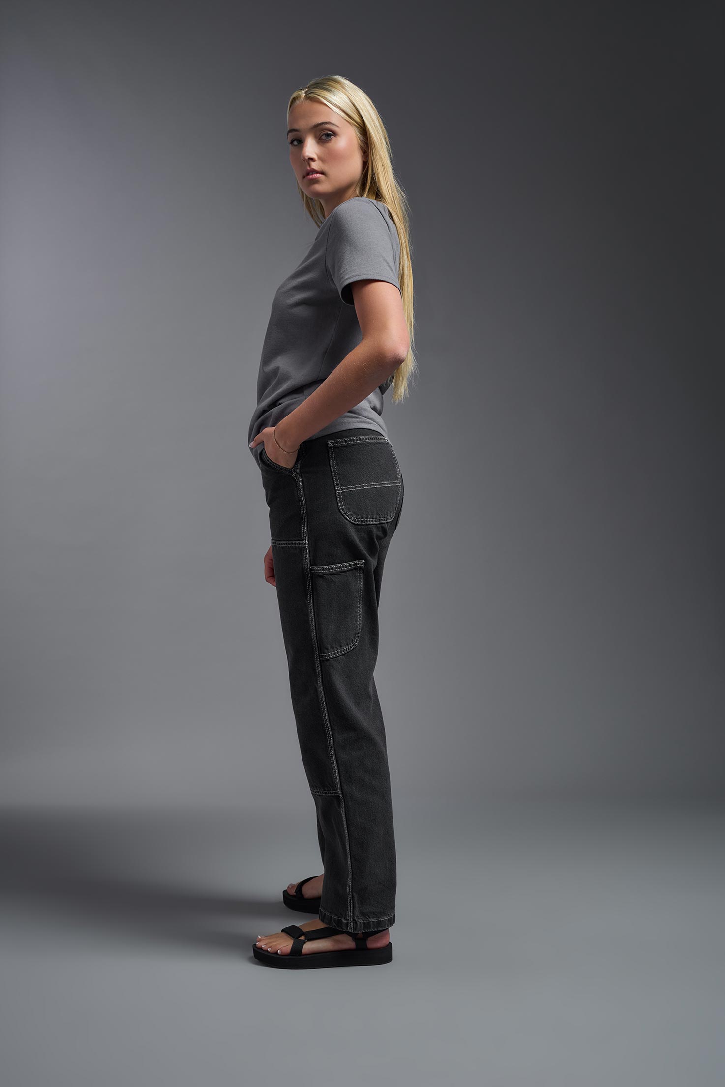 A female model wearing the unisex Imagen Short Sleeve Crewneck T-Shirt in the color carbon, made from 50% U.S. Grown Regenerative Cotton and 50% Recycled Polyester, showing the Relaxed Fit from a side view. This sustainable shirt is Made in the USA. #carbon