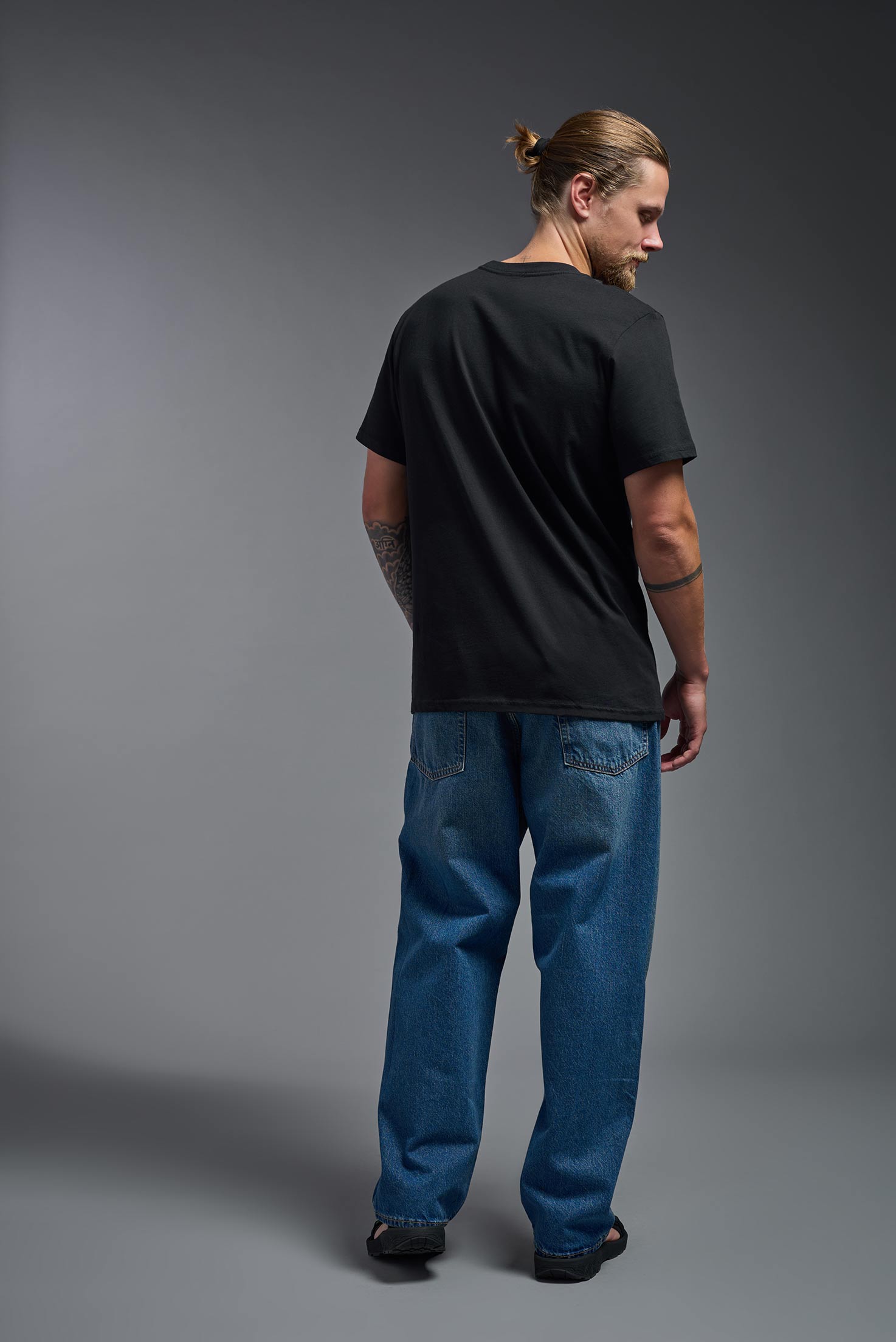 A male model wearing the unisex Organic Soft Cotton T-shirt in the color black, made in the USA from 100% GOTS certified organic cotton from a back view. This shirt is unisex, made in the USA, and perfect for bulk screen printing and decorating. #black