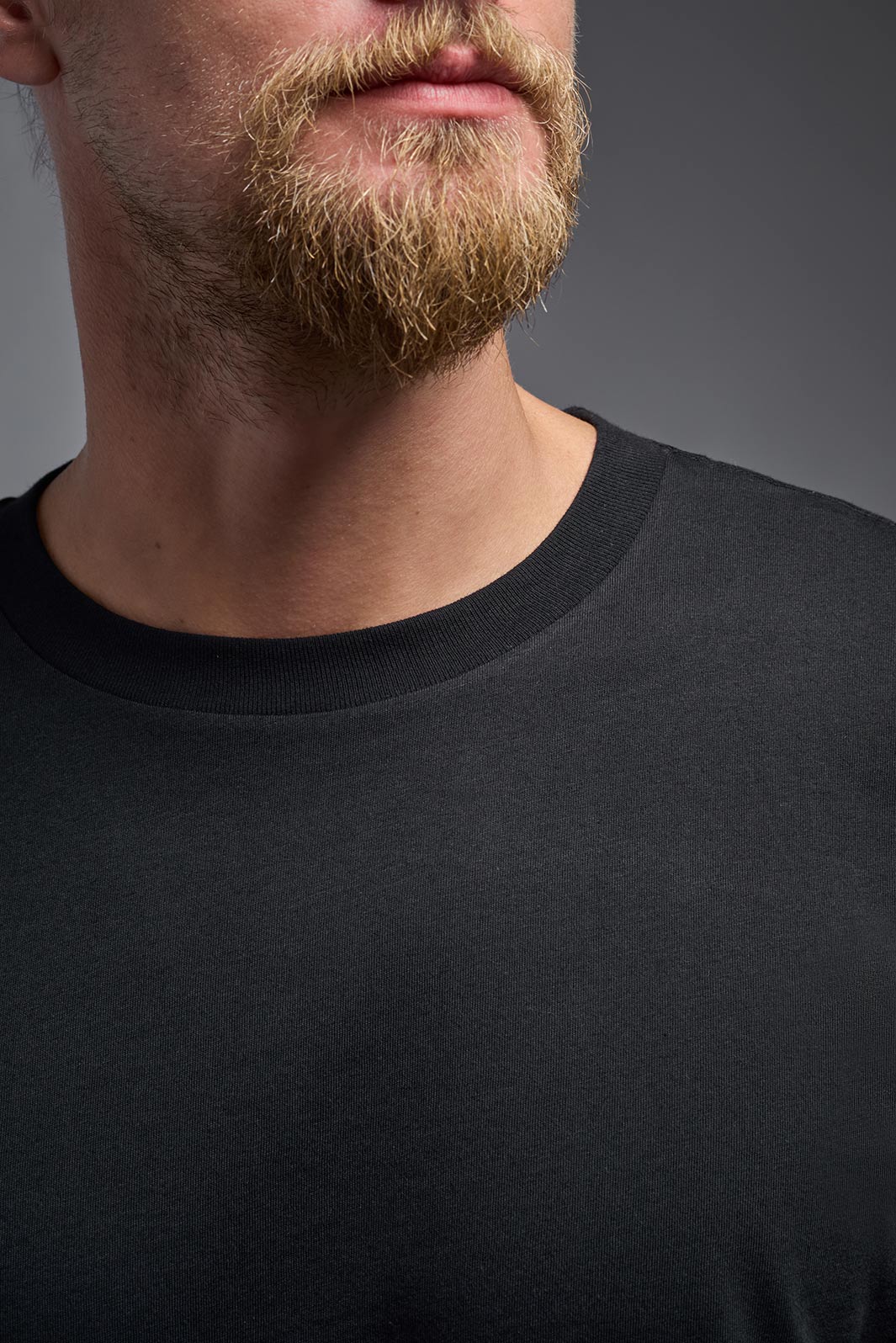 A male model wearing the unisex Organic Soft Cotton T-shirt in the color black, made in the USA from 100% GOTS certified organic cotton from a front close-up collar detail view. #black