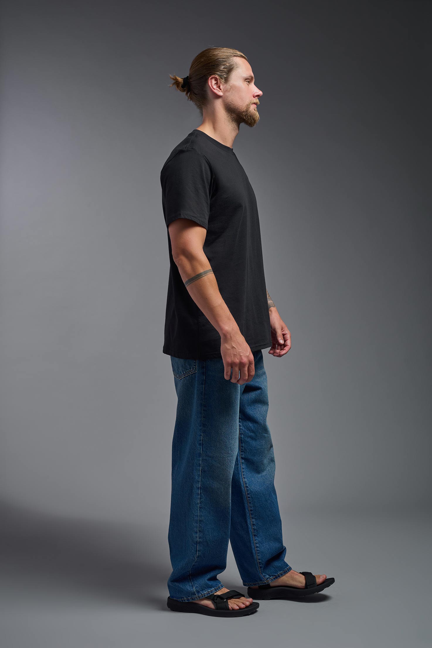 A male model wearing the unisex Organic Soft Cotton T-shirt in the color black, made in the USA from 100% GOTS certified organic cotton from a side view. This shirt is unisex, made in the USA, and perfect for bulk screen printing and decorating. #black