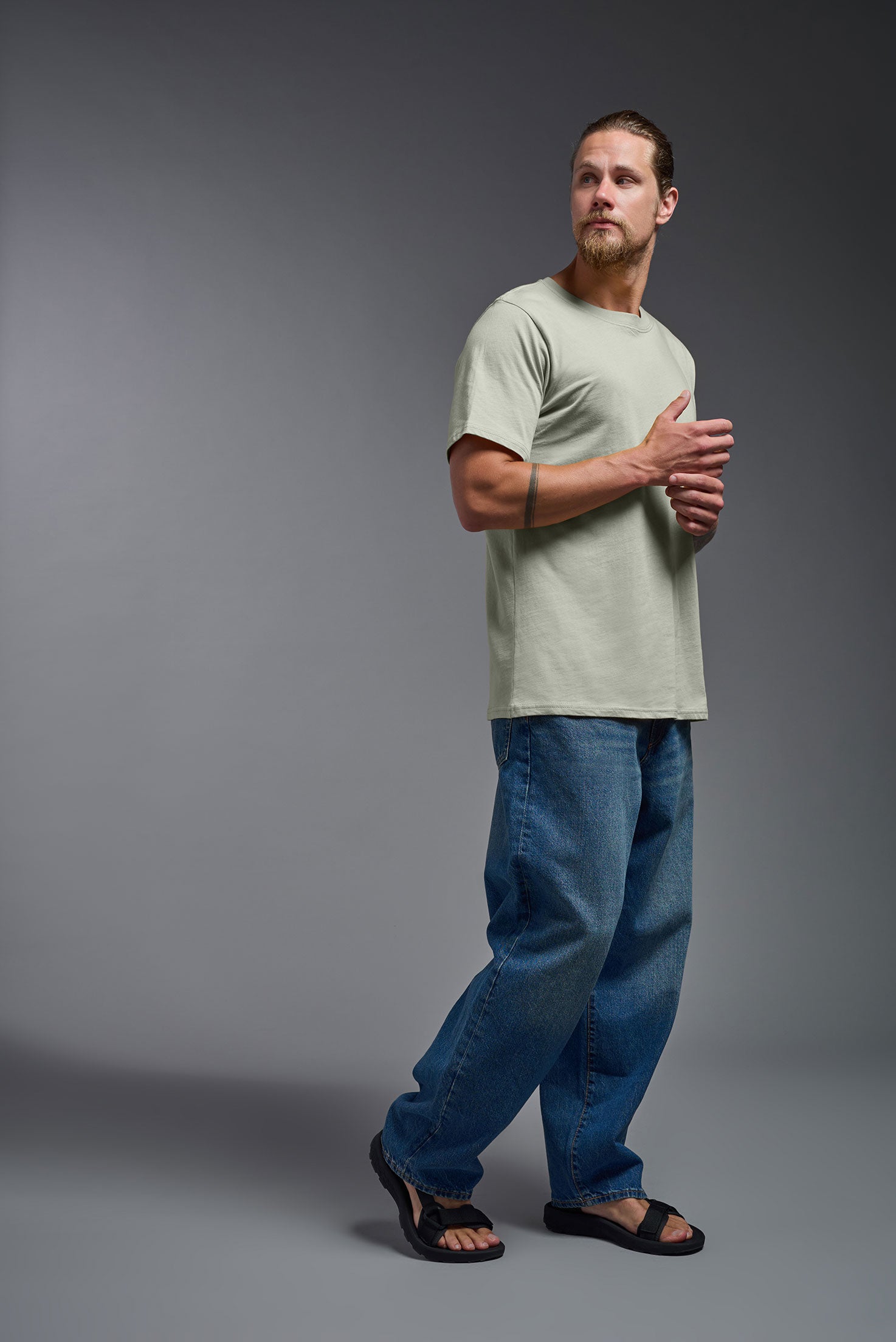 A male model wearing the unisex Organic Soft Cotton T-shirt in the color sage green, made in the USA from 100% GOTS certified organic cotton from a side view. This shirt is unisex, made in the USA, and perfect for bulk screen printing and decorating. #sage