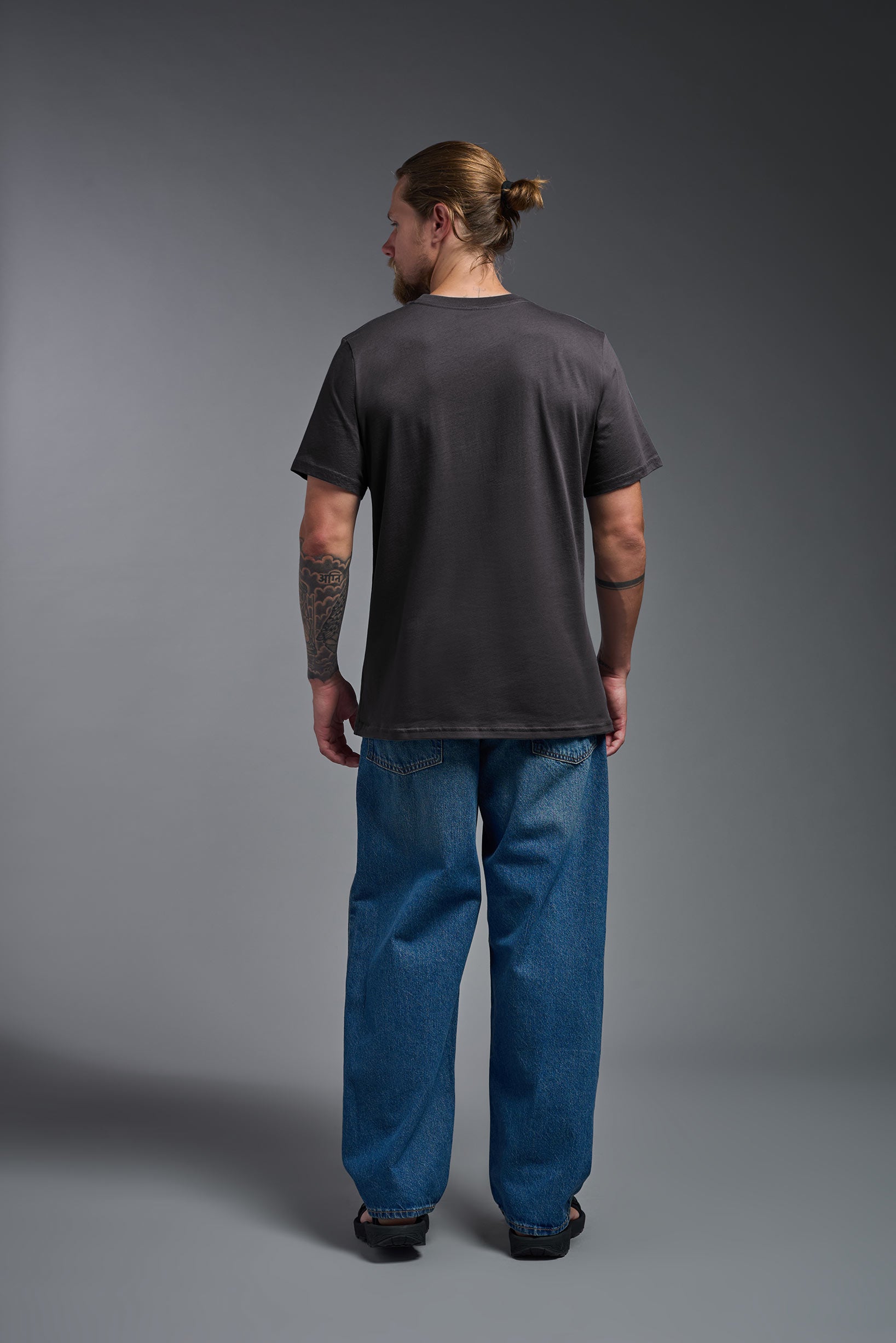 A male model wearing the unisex Organic Soft Cotton T-shirt in the color carbon, made from 100% GOTS certified organic cotton from a full body back view. This shirt is unisex, made in the USA, and perfect for bulk screen printing and decorating. #carbon