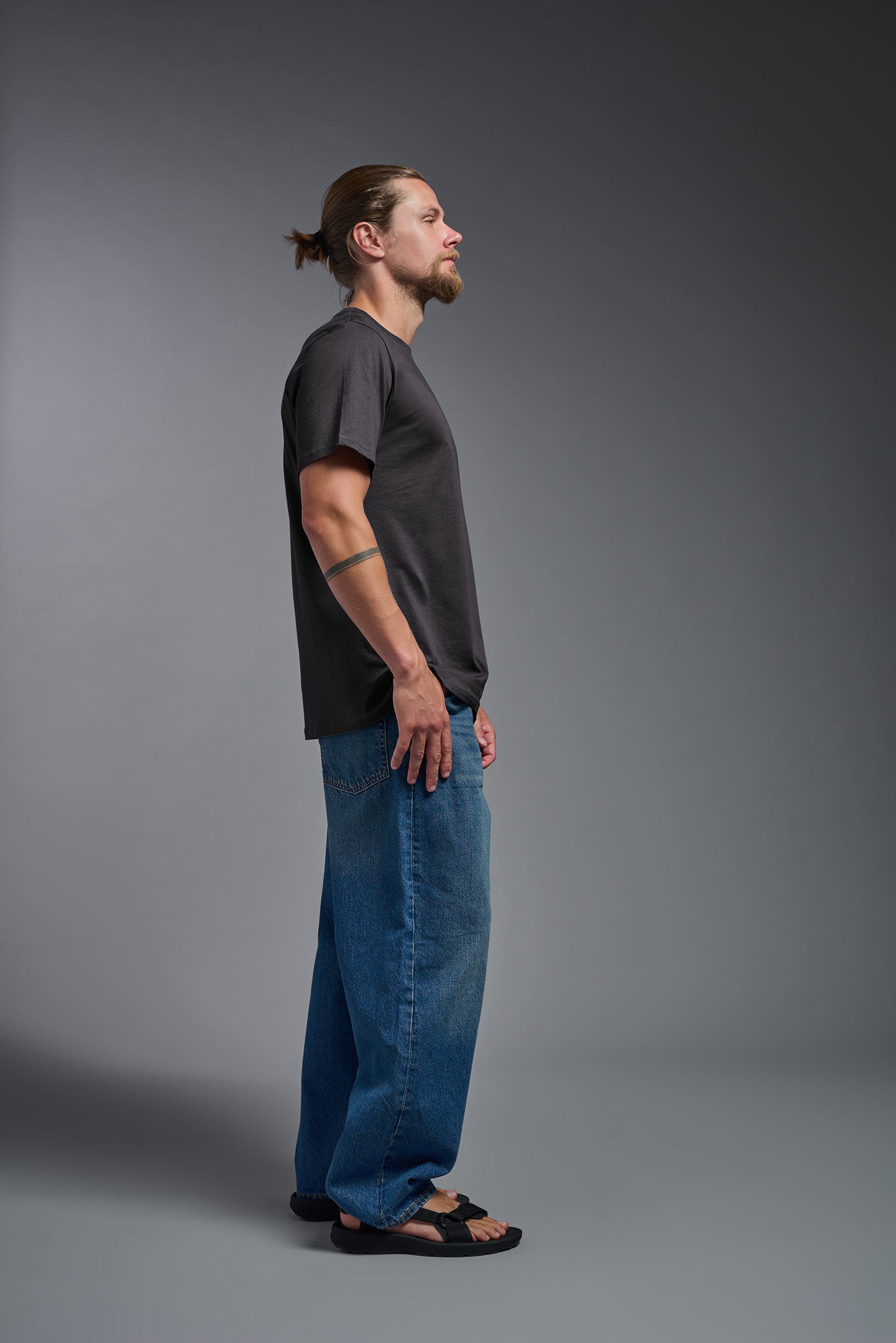 A male model wearing the unisex Organic Soft Cotton T-shirt in the color carbon, made from 100% GOTS certified organic cotton from a full body side view. This shirt is unisex, made in the USA, and perfect for bulk screen printing and decorating. #carbon