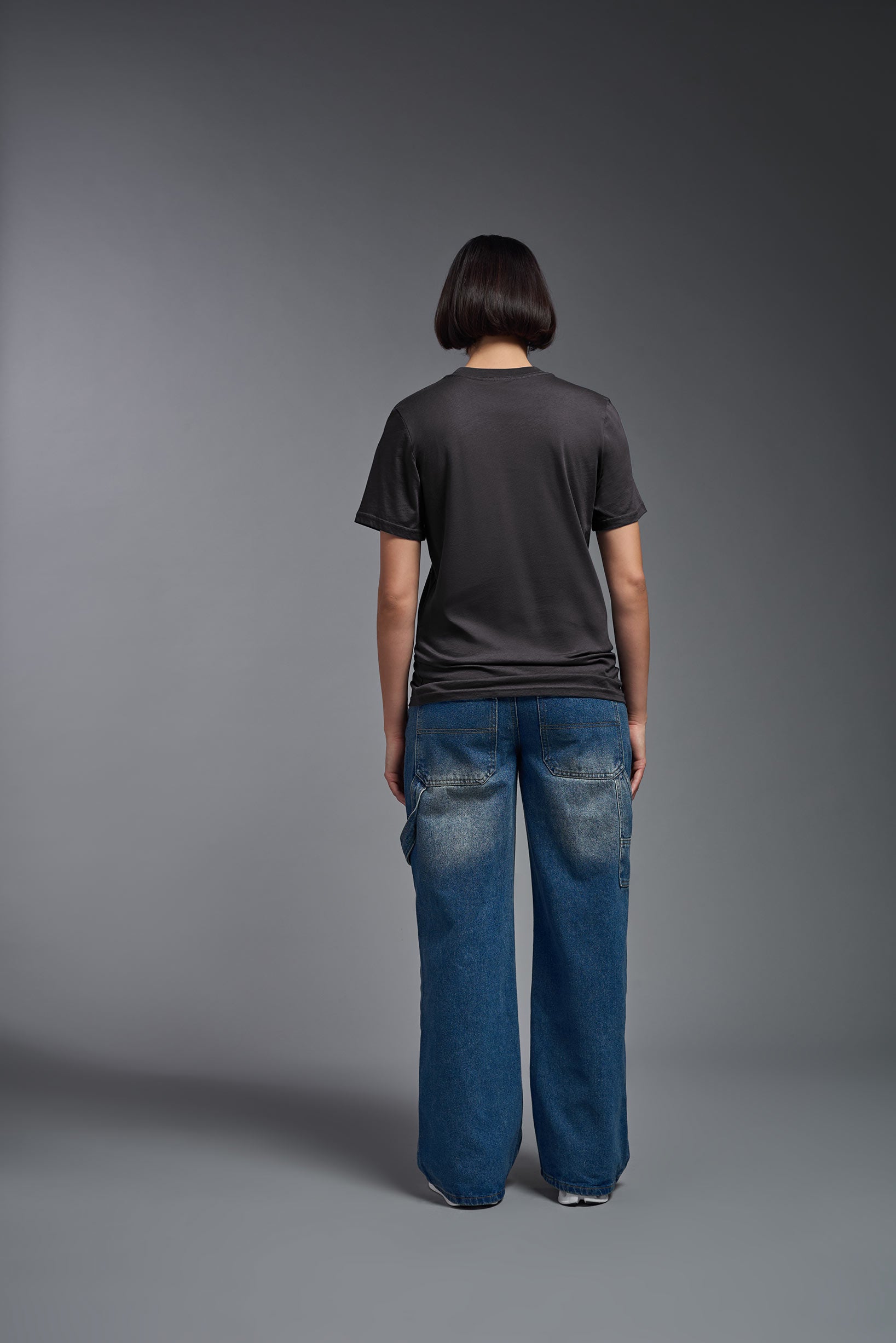 A female model wearing the unisex Organic Soft Cotton T-shirt in the color carbon, made from 100% GOTS certified organic cotton from a full body back view. This shirt is unisex, made in the USA, and perfect for bulk screen printing and decorating. #carbon