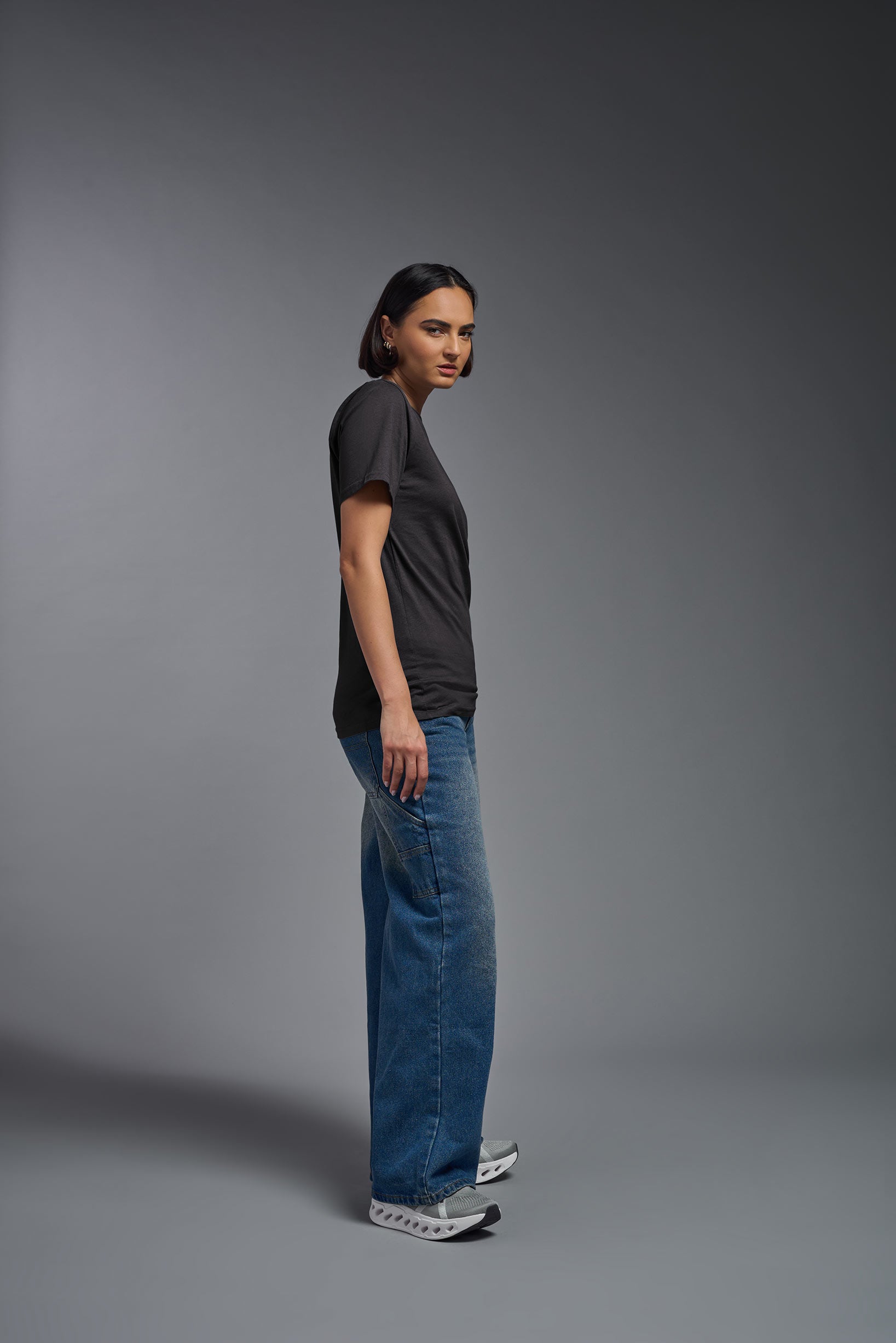 A female model wearing the unisex Organic Soft Cotton T-shirt in the color carbon, made from 100% GOTS certified organic cotton from a full body side view. This shirt is unisex, made in the USA, and perfect for bulk screen printing and decorating. #carbon