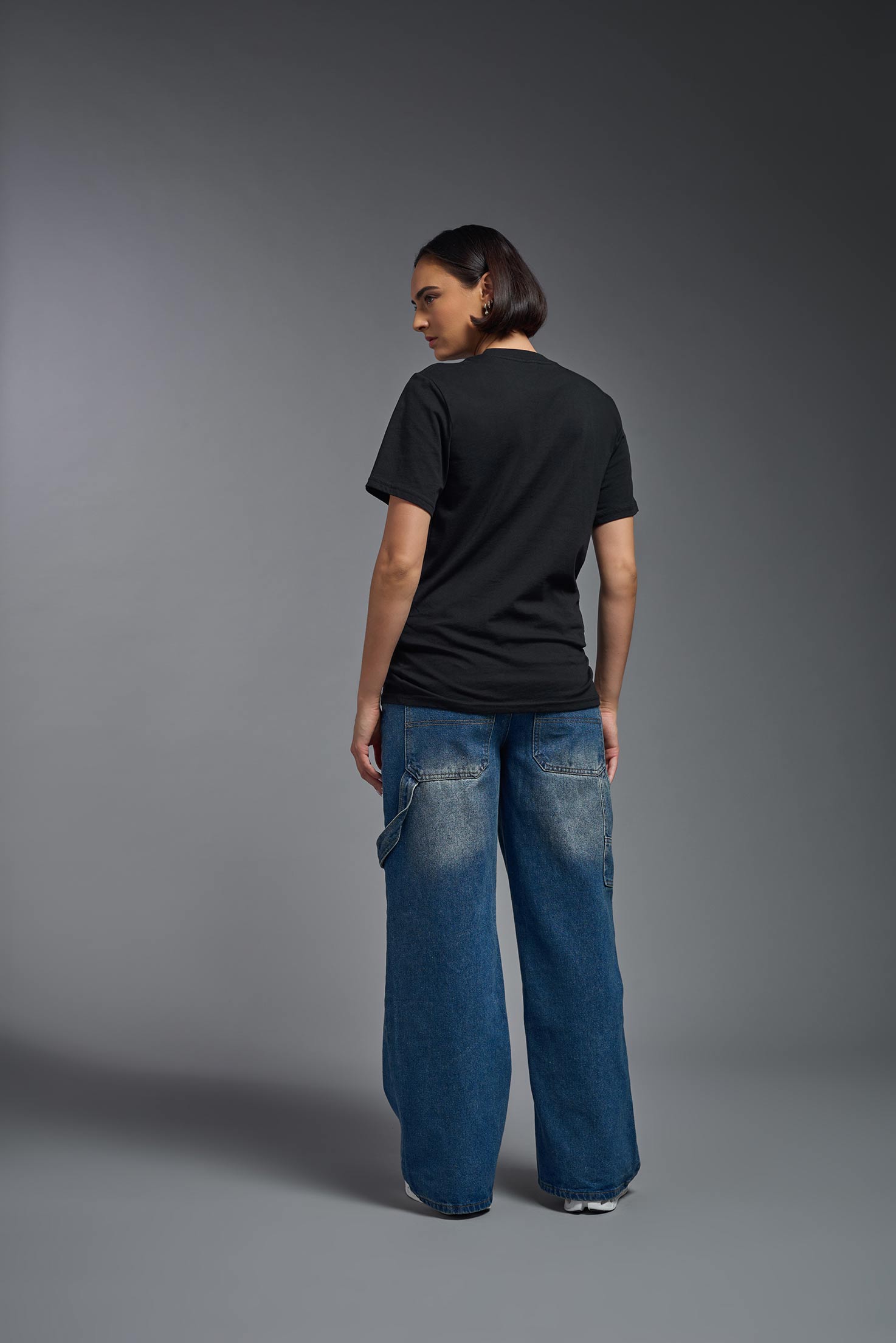 A female model wearing the unisex Organic Soft Cotton T-shirt in the color black, made in the USA from 100% GOTS certified organic cotton from a back view. This shirt is unisex, made in the USA, and perfect for bulk screen printing and decorating. #black