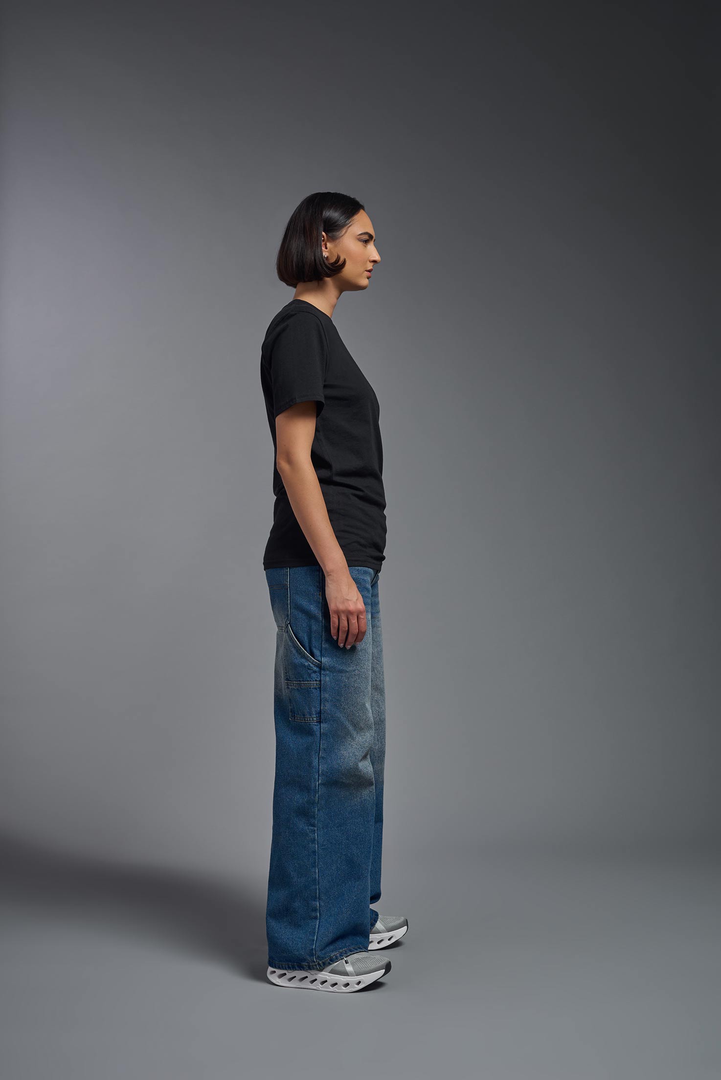 A female model wearing the unisex Organic Soft Cotton T-shirt in the color black, made in the USA from 100% GOTS certified organic cotton from a side view. This shirt is unisex, made in the USA, and perfect for bulk screen printing and decorating. #black