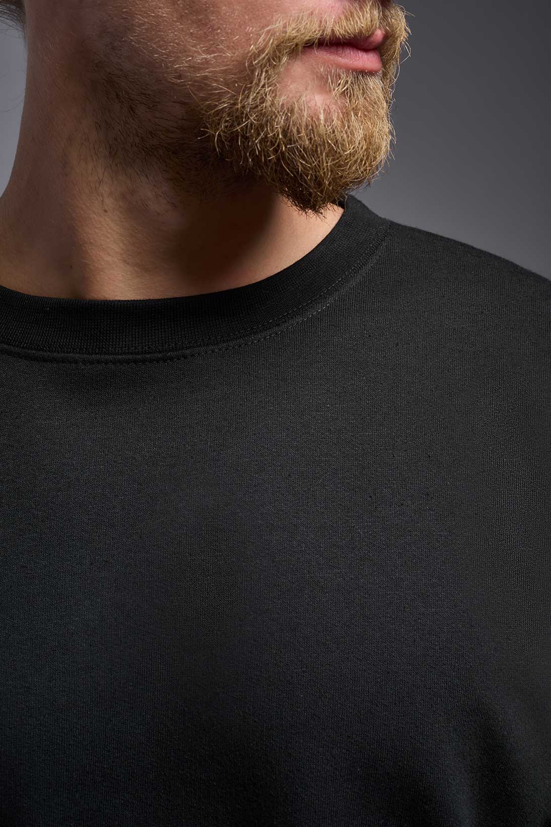 A male model wearing the unisex Americana 3-End Fleece Crewneck Sweatshirt in the color black, made from 75% combed ring-spun USA-grown cotton and 25% polyester, showing the relaxed fit from a close-up collar view. This premium fleece hoodie is Made in the USA. #black