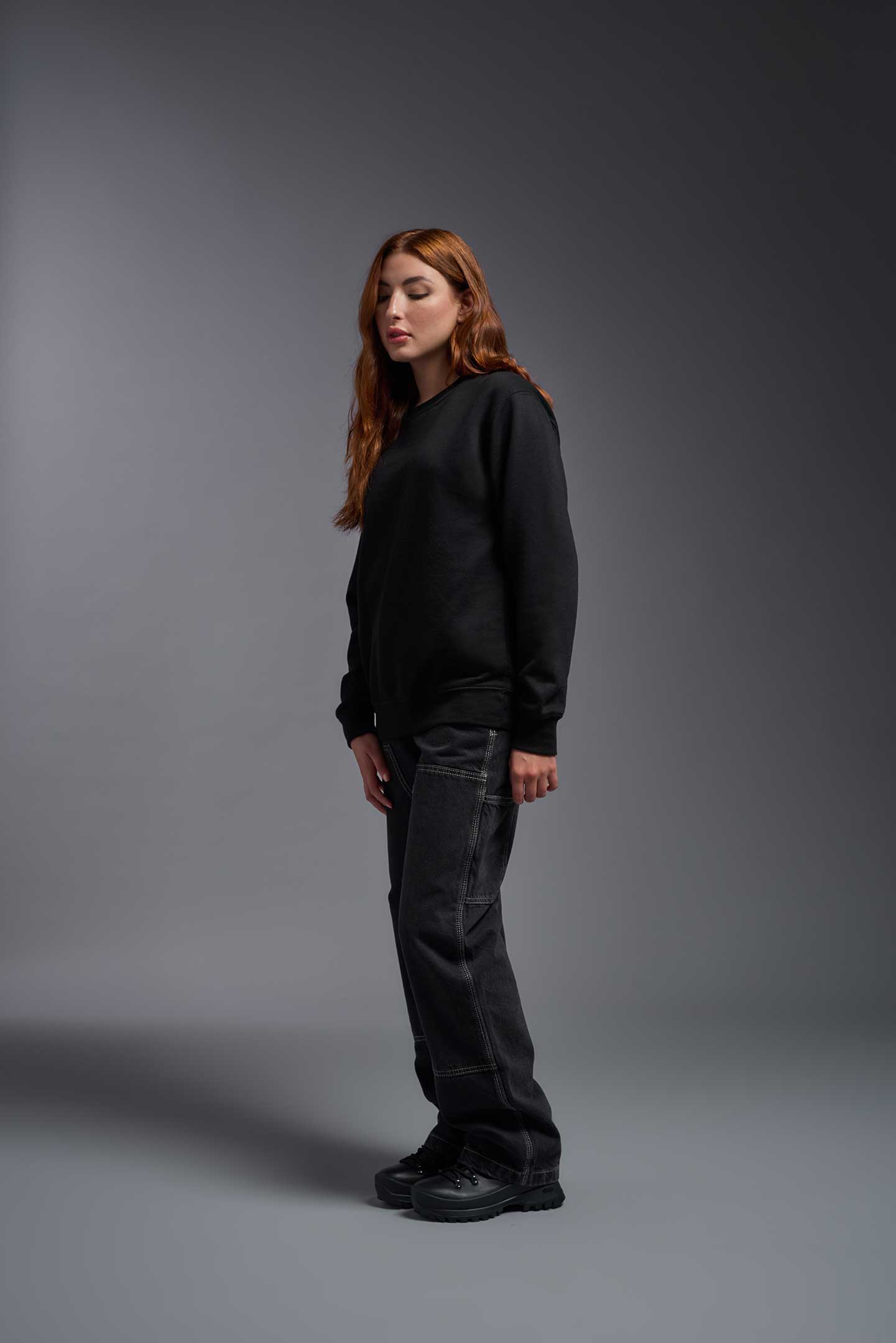 A female model wearing the unisex Americana 3-End Fleece Crewneck Sweatshirt in the color black, made from 75% combed ring-spun USA-grown cotton and 25% polyester, showing the relaxed fit from a side view. This premium fleece hoodie is Made in the USA. #black