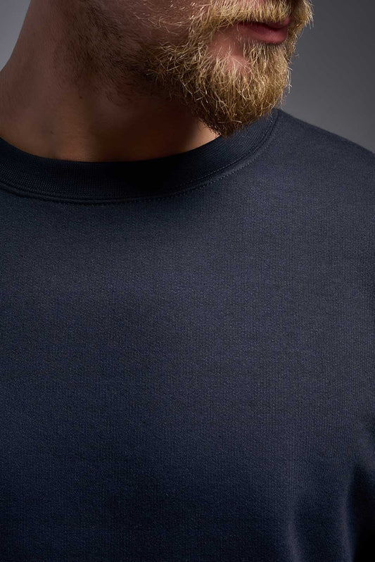 A male model wearing the unisex Americana 3-End Fleece Crewneck Sweatshirt in the color navy, made from 75% combed ring-spun USA-grown cotton and 25% polyester, showing the relaxed fit from a close-up collar view. This premium fleece hoodie is Made in the USA. #navy