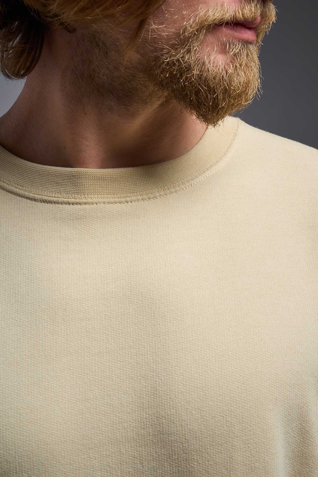 A male model wearing the unisex Americana 3-End Fleece Crewneck Sweatshirt in the color sand, made from 75% combed ring-spun USA-grown cotton and 25% polyester, showing the relaxed fit from a close-up collar view. This premium fleece hoodie is Made in the USA. #sand