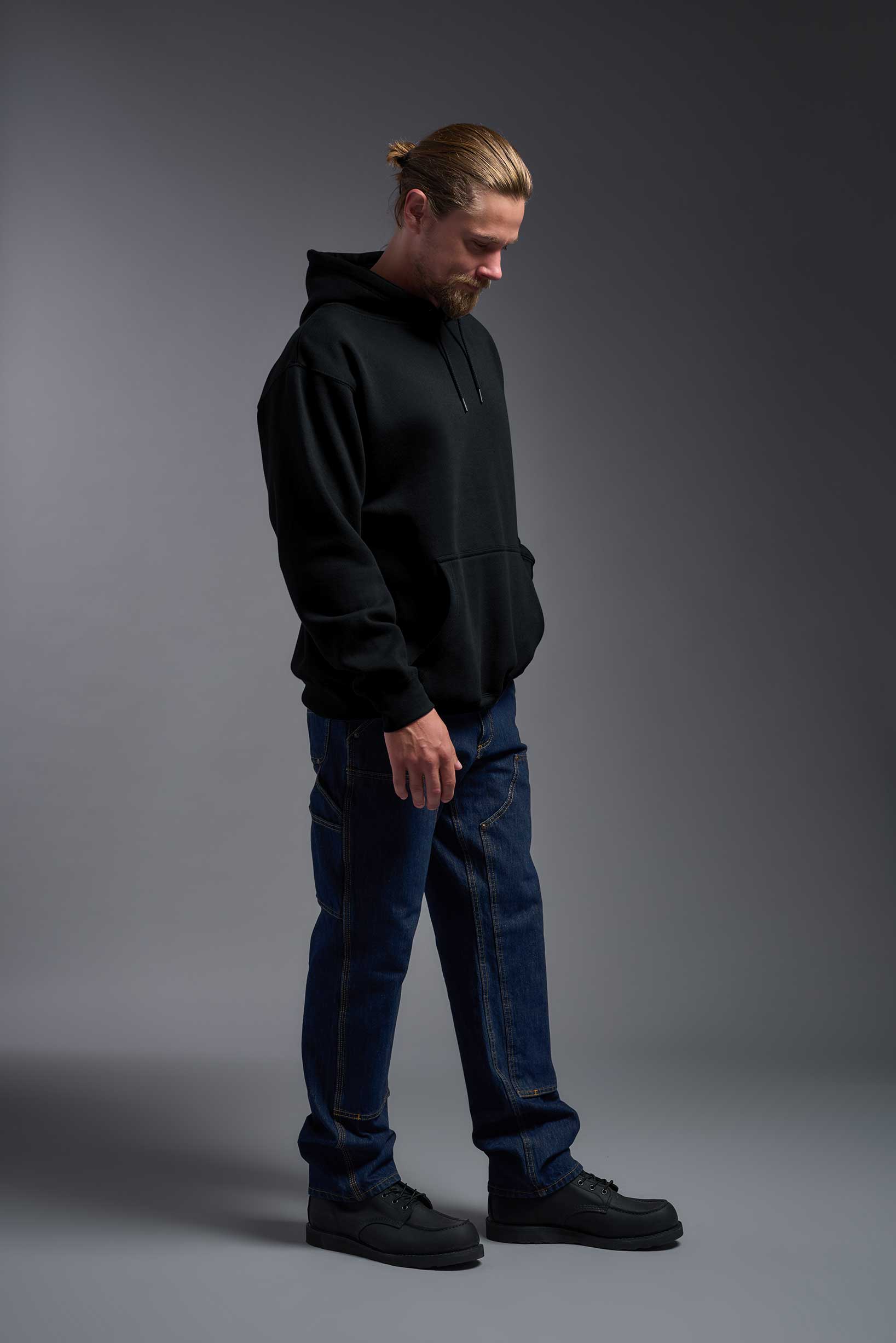 A male model wearing the unisex Americana 3-End Fleece Pullover Hoodie in the color black, made from 75% combed ring-spun USA-grown cotton and 25% polyester, showing the relaxed fit from a side view. This premium fleece hoodie is Made in the USA. #black