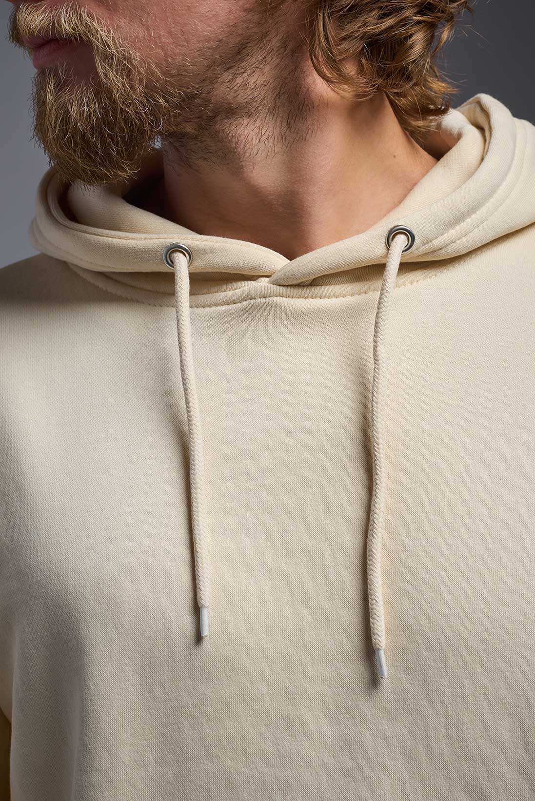 A male model wearing the unisex Americana 3-End Fleece Pullover Hoodie in the color sand, made from 75% combed ring-spun USA-grown cotton and 25% polyester, showing the relaxed fit from a close-up collar view. This premium fleece hoodie is Made in the USA. #sand