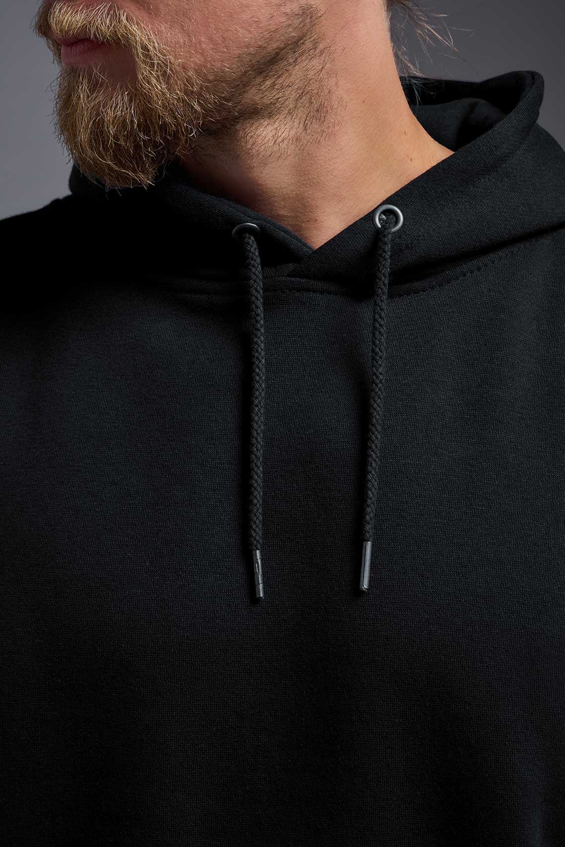 A male model wearing the unisex Americana 3-End Fleece Pullover Hoodie in the color black, made from 75% combed ring-spun USA-grown cotton and 25% polyester, showing the relaxed fit from a close-up collar view. This premium fleece hoodie is Made in the USA. #black