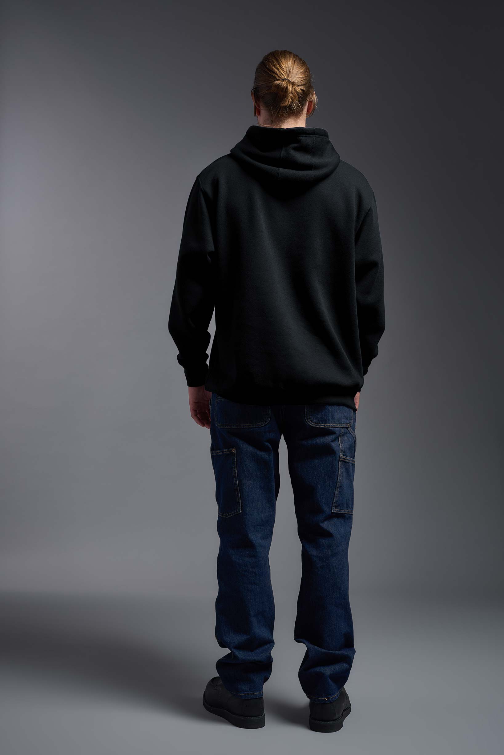 A male model wearing the unisex Americana 3-End Fleece Pullover Hoodie in the color black, made from 75% combed ring-spun USA-grown cotton and 25% polyester, showing the relaxed fit from a back view. This premium fleece hoodie is Made in the USA. #black