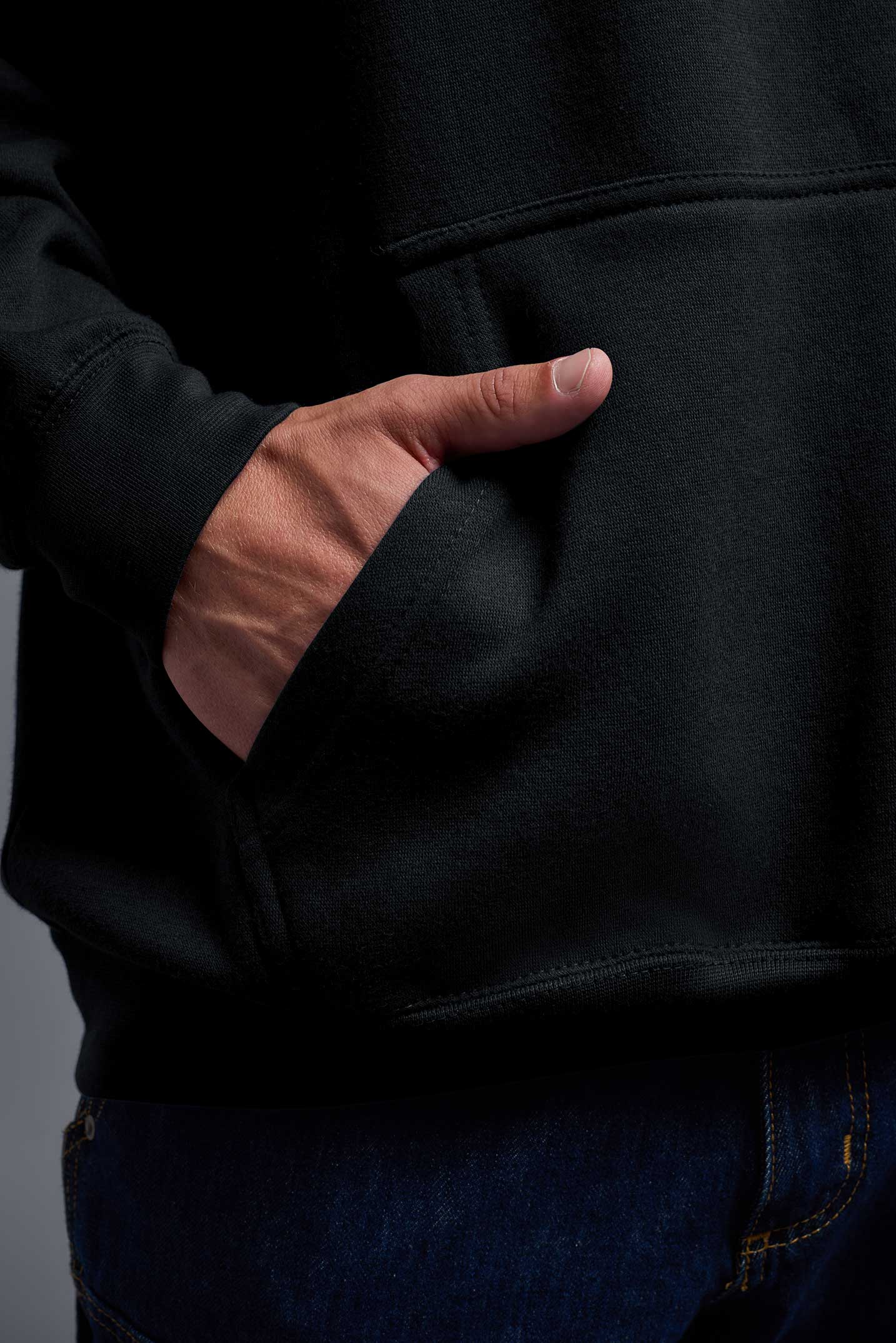 A male model wearing the unisex Americana 3-End Fleece Pullover Hoodie in the color black, made from 75% combed ring-spun USA-grown cotton and 25% polyester, showing the relaxed fit from a close-up pocket view. This premium fleece hoodie is Made in the USA. #black