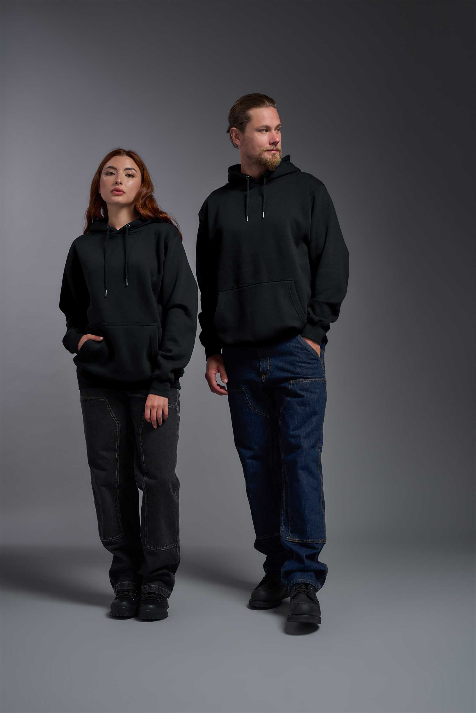 A male and female model wearing the unisex Americana 3-End Fleece Pullover Hoodie in the color black, made from 75% combed ring-spun USA-grown cotton and 25% polyester, showing the relaxed fit from a front view. This premium fleece hoodie is Made in the USA. #black