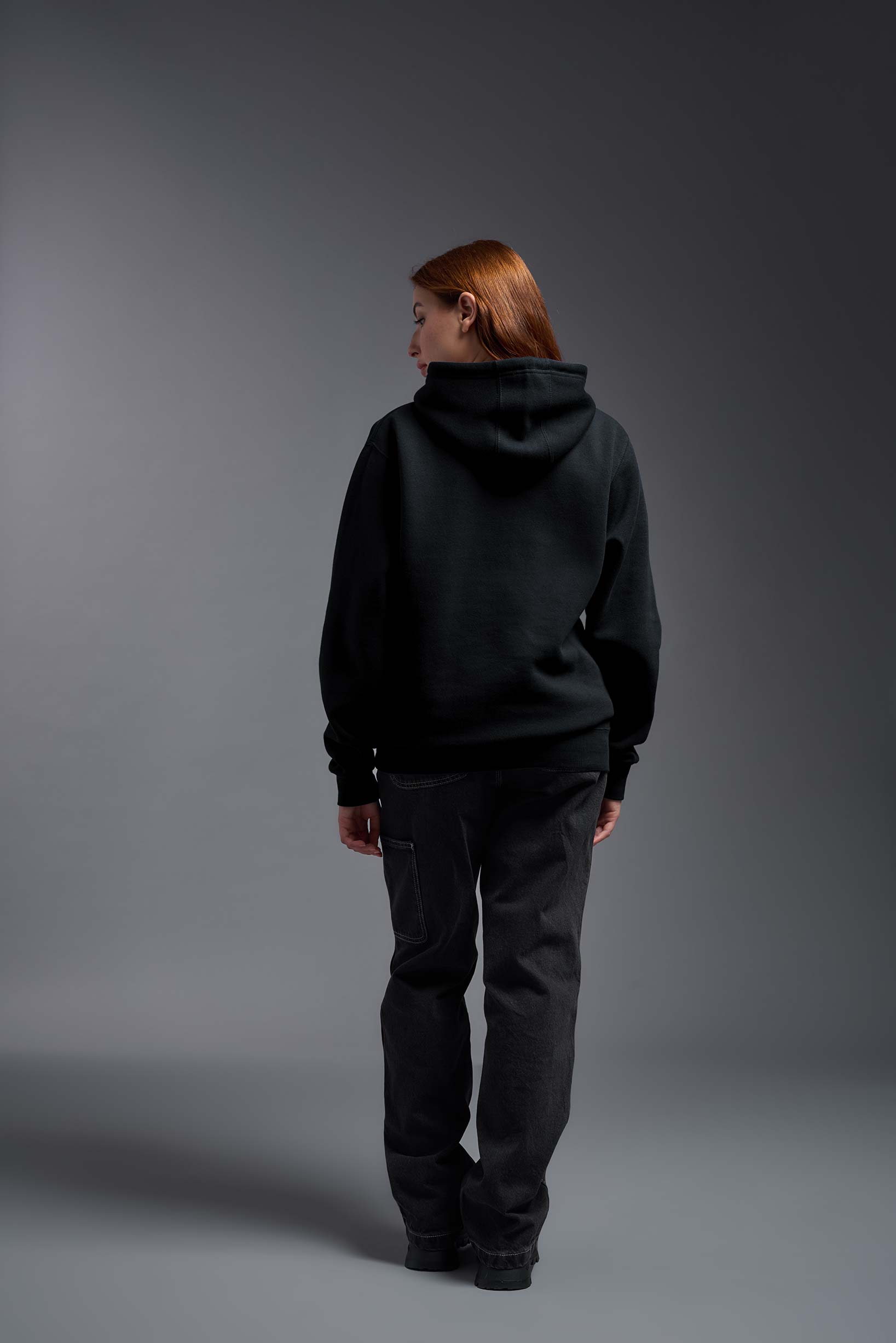 A female model wearing the unisex Americana 3-End Fleece Pullover Hoodie in the color black, made from 75% combed ring-spun USA-grown cotton and 25% polyester, showing the relaxed fit from a back view. This premium fleece hoodie is Made in the USA. #black