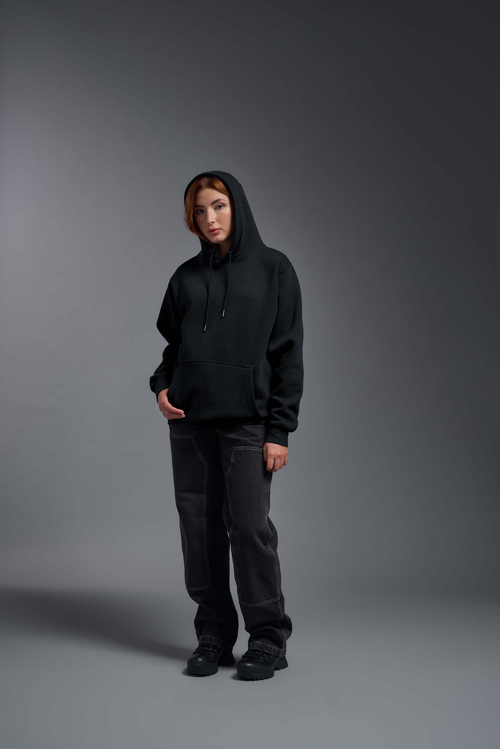 A female model wearing the unisex Americana 3-End Fleece Pullover Hoodie in the color black, made from 75% combed ring-spun USA-grown cotton and 25% polyester, showing the relaxed fit from a front view with hood up. This premium fleece hoodie is Made in the USA. #black