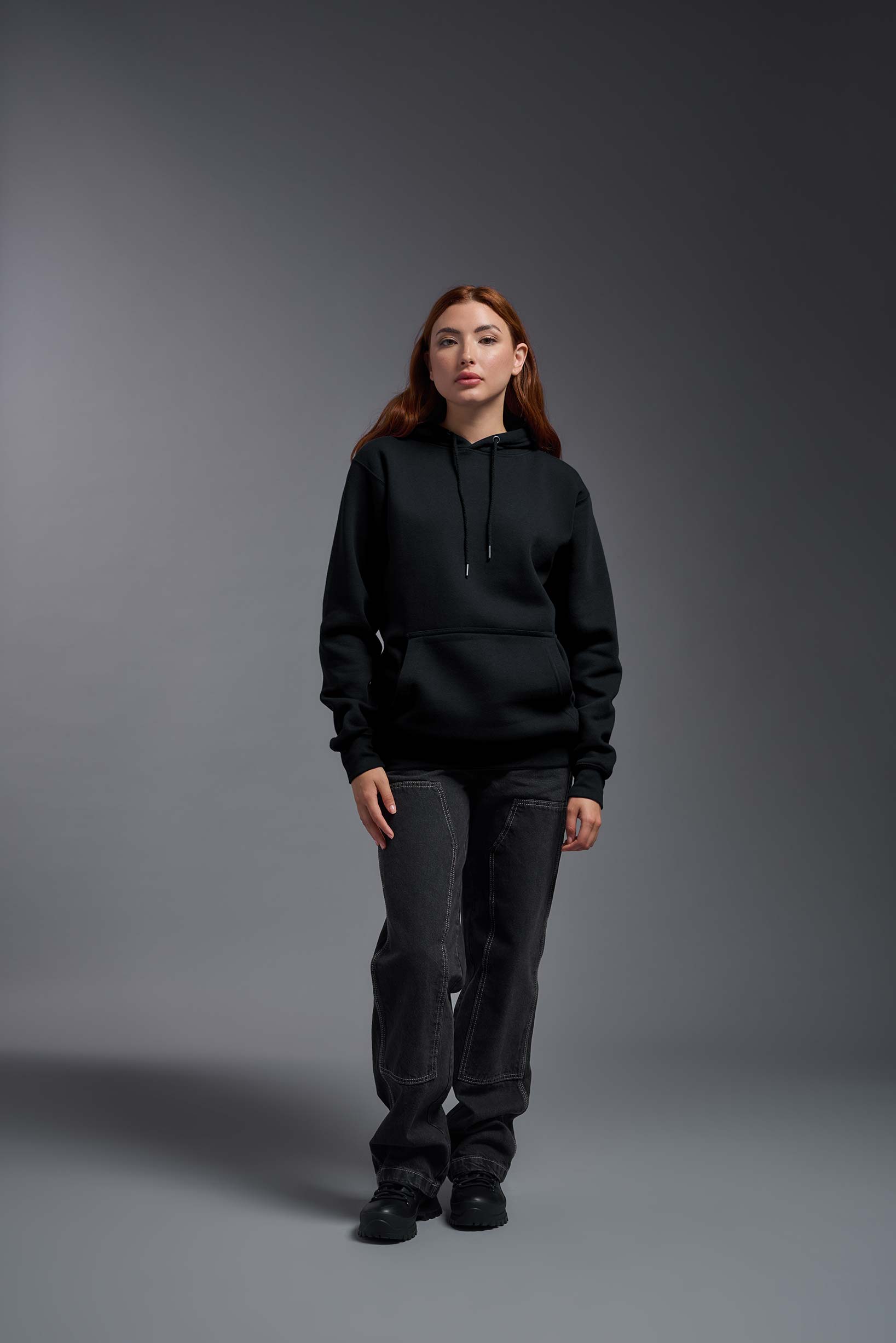A female model wearing the unisex Americana 3-End Fleece Pullover Hoodie in the color black, made from 75% combed ring-spun USA-grown cotton and 25% polyester, showing the relaxed fit from a front view. This premium fleece hoodie is Made in the USA. #black