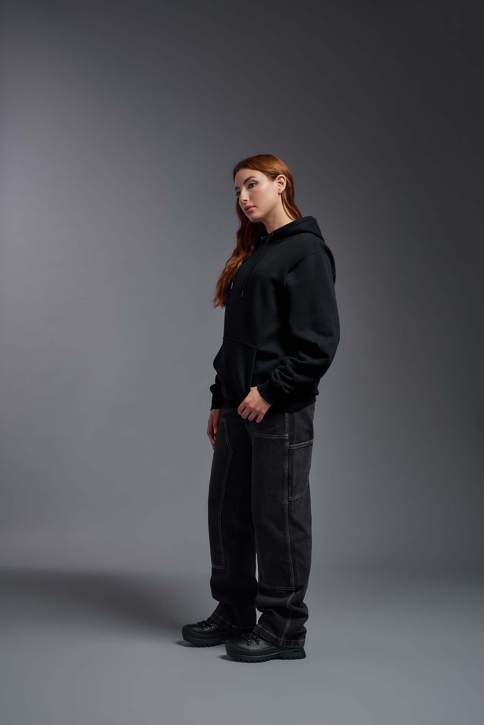 A female model wearing the unisex Americana 3-End Fleece Pullover Hoodie in the color black, made from 75% combed ring-spun USA-grown cotton and 25% polyester, showing the relaxed fit from a side view. This premium fleece hoodie is Made in the USA. #black
