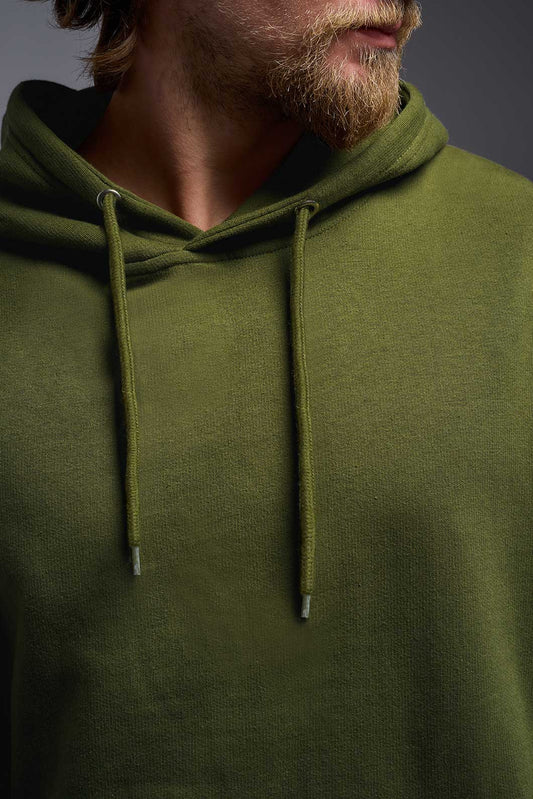 A male model wearing the unisex Americana 3-End Fleece Pullover Hoodie in the color military green, made from 75% combed ring-spun USA-grown cotton and 25% polyester, showing the relaxed fit from a close-up collar view. This premium fleece hoodie is Made in the USA. #military-green