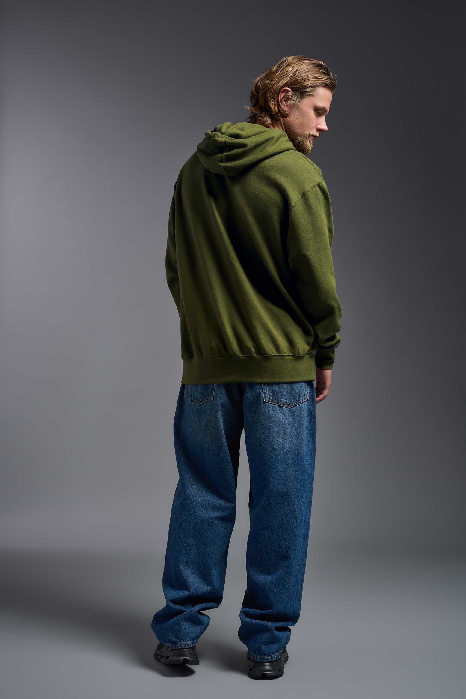 A male model wearing the unisex Americana 3-End Fleece Pullover Hoodie in the color military green, made from 75% combed ring-spun USA-grown cotton and 25% polyester, showing the relaxed fit from a back view. This premium fleece hoodie is Made in the USA. #military-green