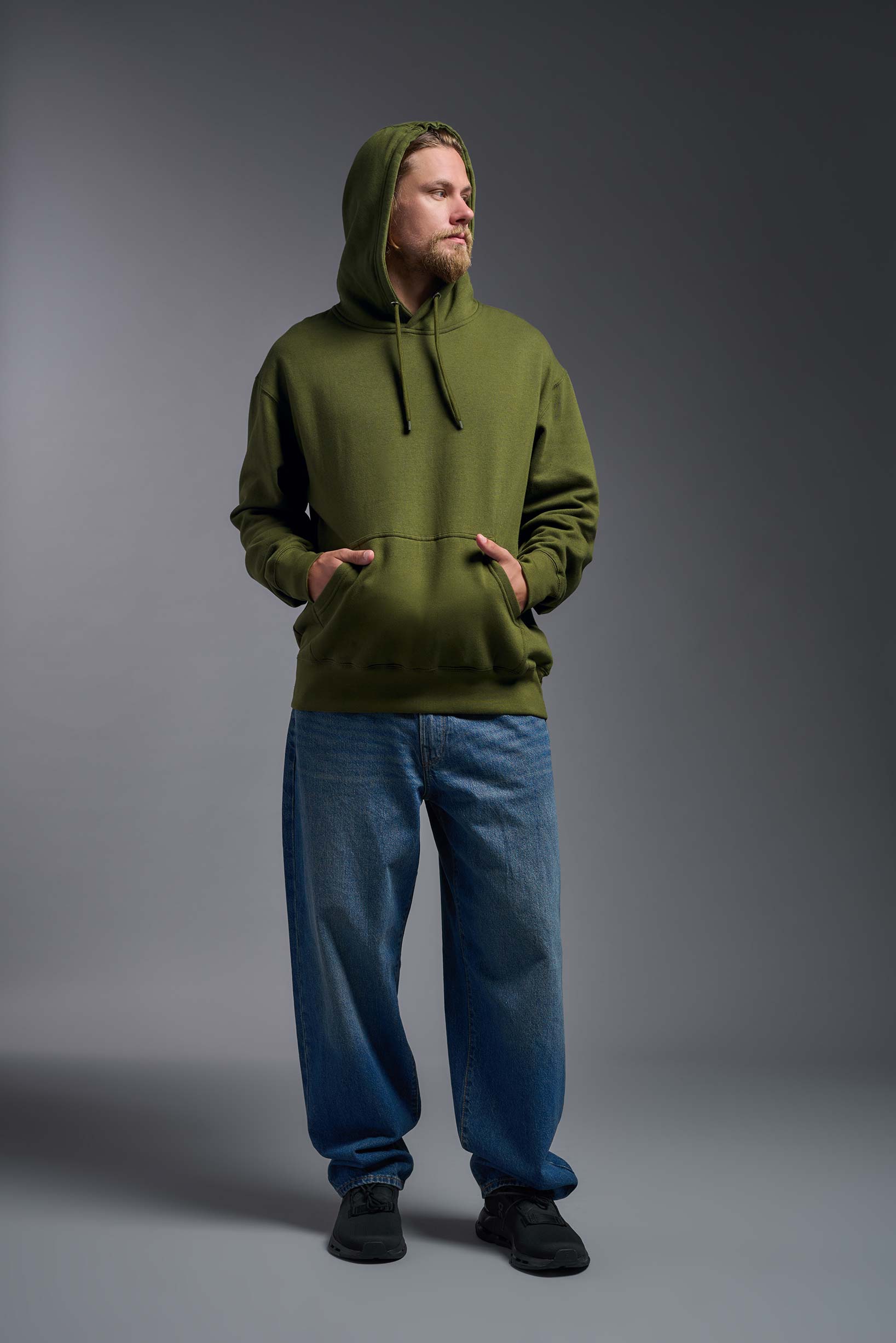 A male model wearing the unisex Americana 3-End Fleece Pullover Hoodie in the color military green, made from 75% combed ring-spun USA-grown cotton and 25% polyester, showing the relaxed fit from a front view with hood up. This premium fleece hoodie is Made in the USA. #military-green