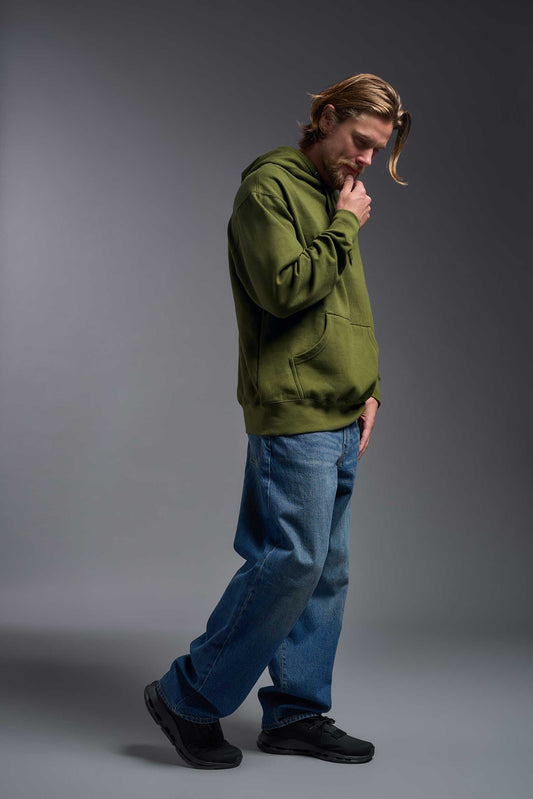 A male model wearing the unisex Americana 3-End Fleece Pullover Hoodie in the color military green, made from 75% combed ring-spun USA-grown cotton and 25% polyester, showing the relaxed fit from a side view. This premium fleece hoodie is Made in the USA. #military-green