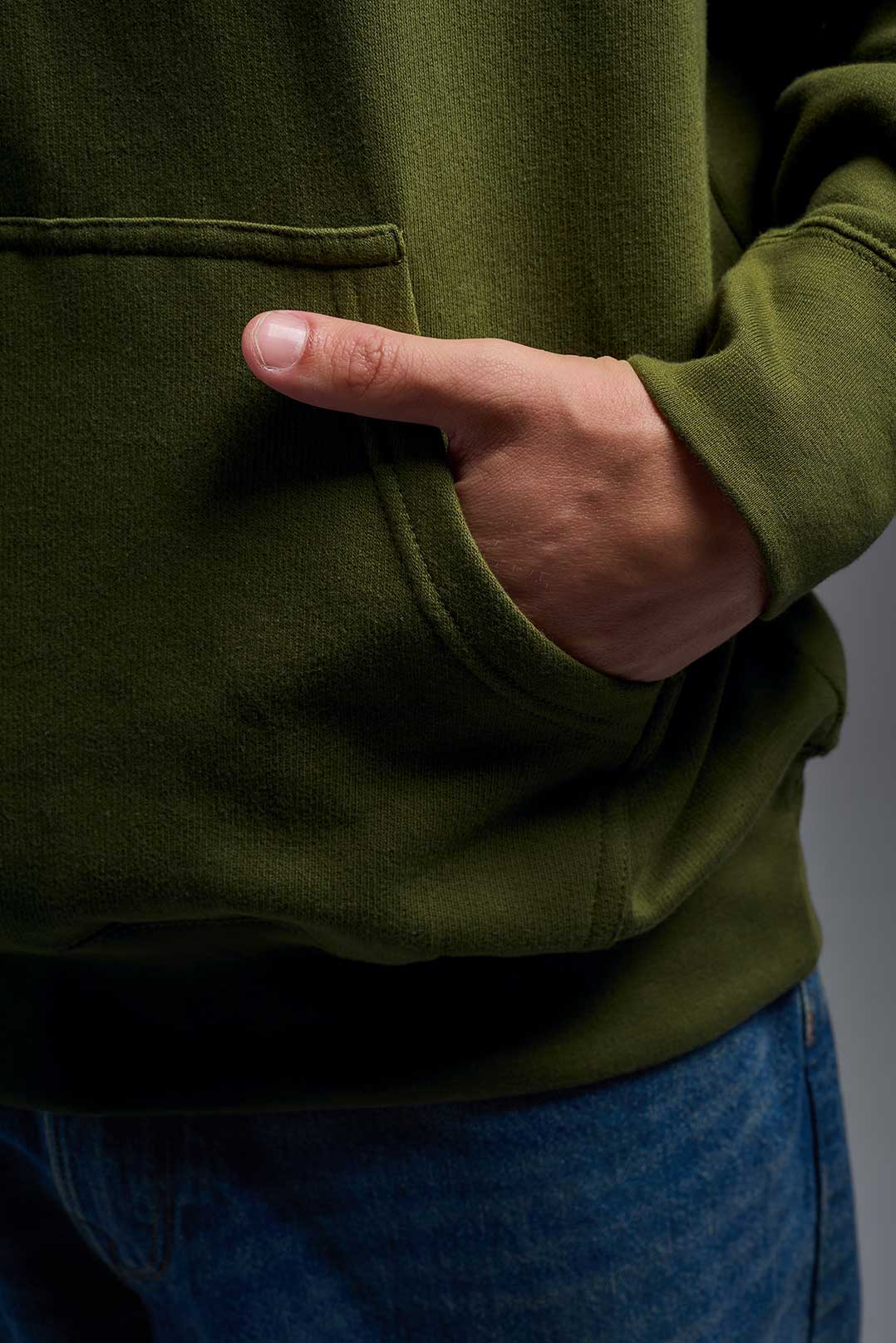 A male model wearing the unisex Americana 3-End Fleece Pullover Hoodie in the color military green, made from 75% combed ring-spun USA-grown cotton and 25% polyester, showing the relaxed fit from a close-up pocket view. This premium fleece hoodie is Made in the USA. #military-green